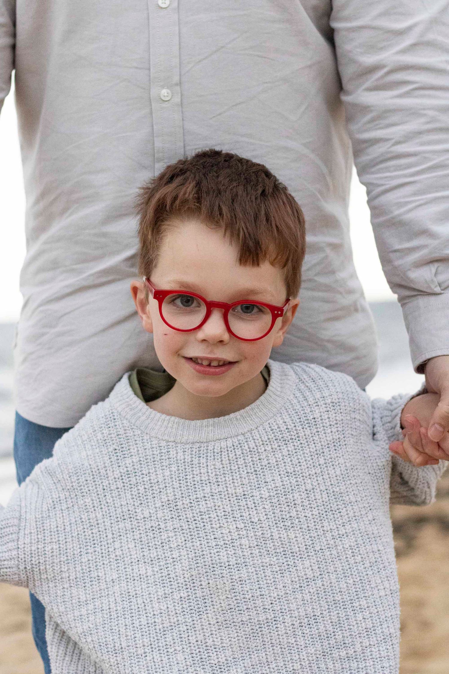 A child wearing a white sweater and red glasses smiles at the camera.