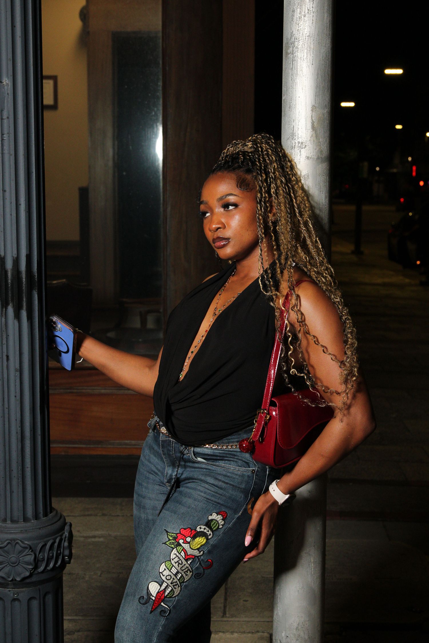 Woman with long braided hair in black top and jeans holding a red purse leaning on a column at night.