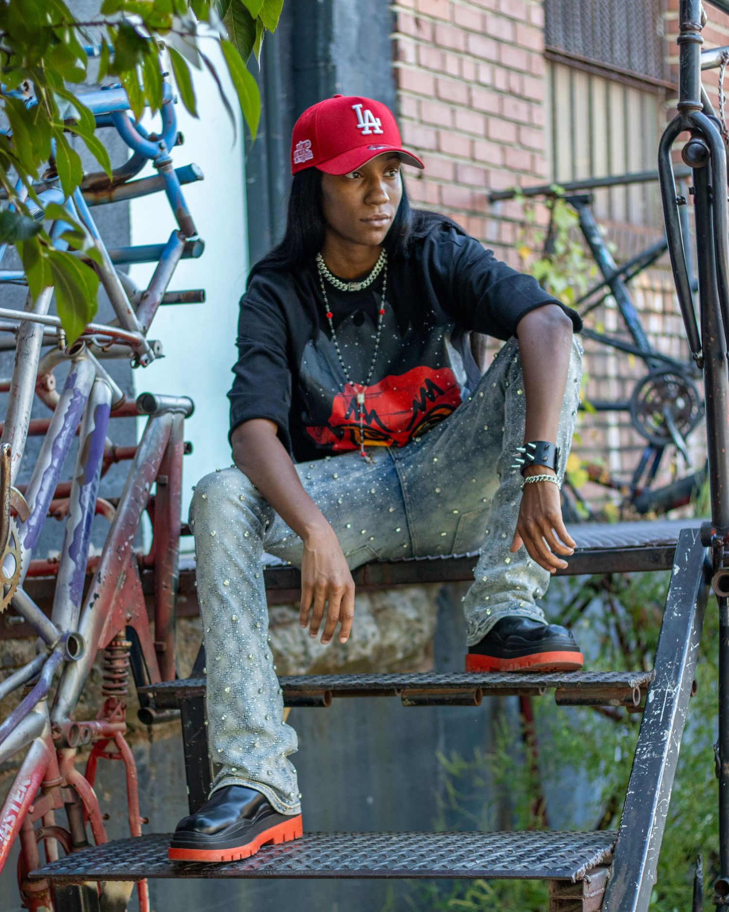 Young man in red LA cap and black hoodie sits on metal stairs outdoors.