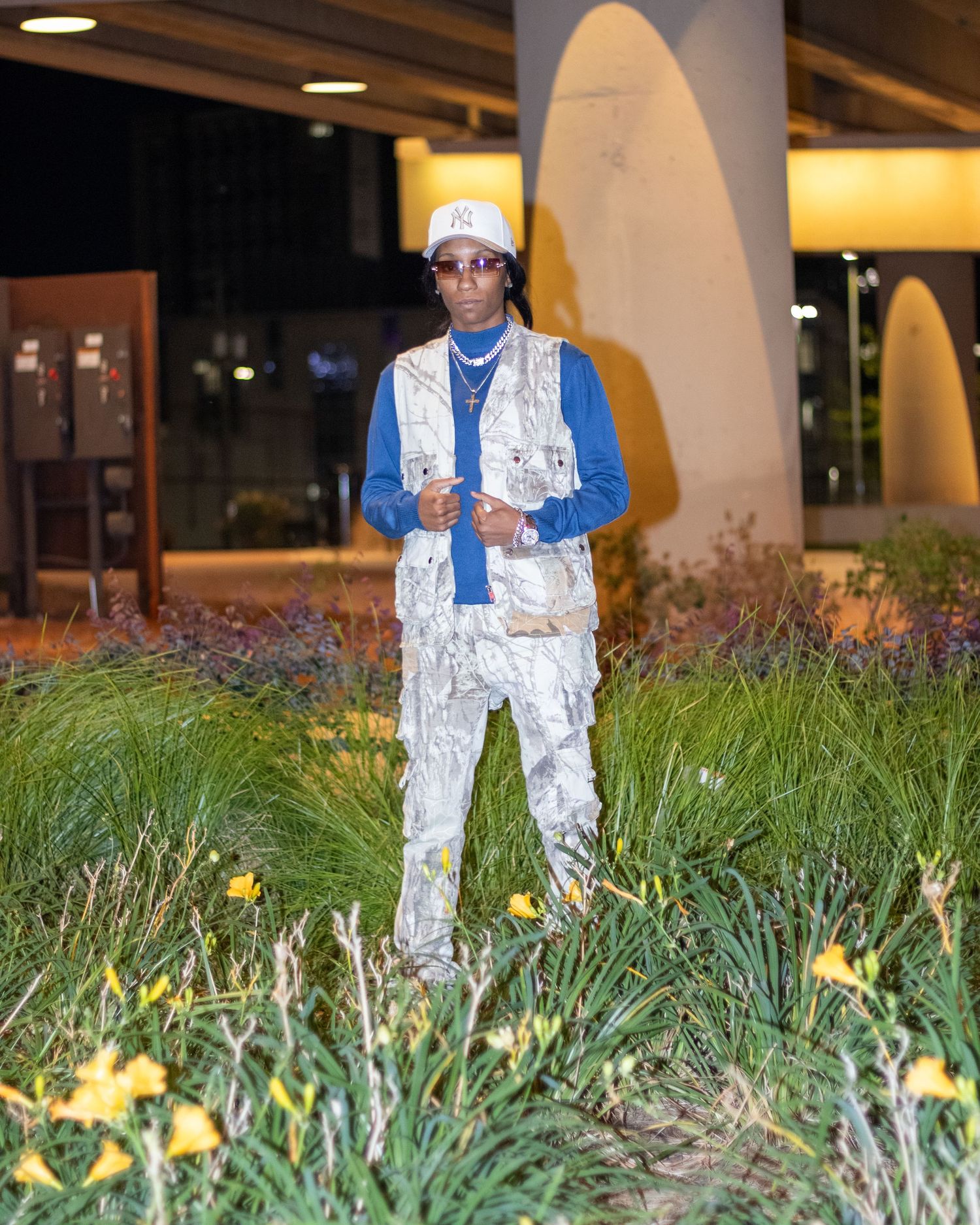 Young man in silver outfit and NY cap standing among flowers at night.