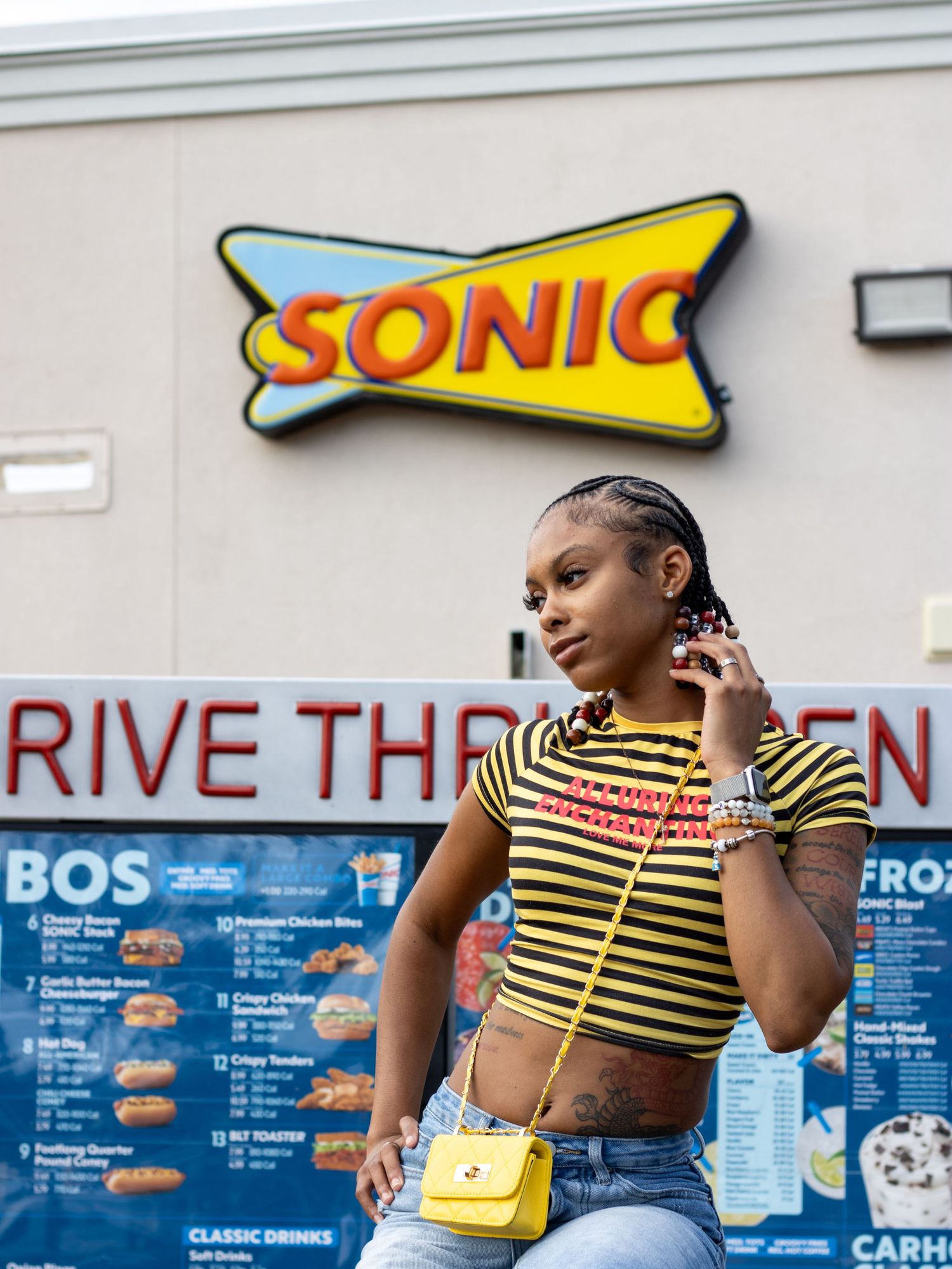 Young woman in striped crop top with yellow purse poses in front of Sonic Drive-In sign.