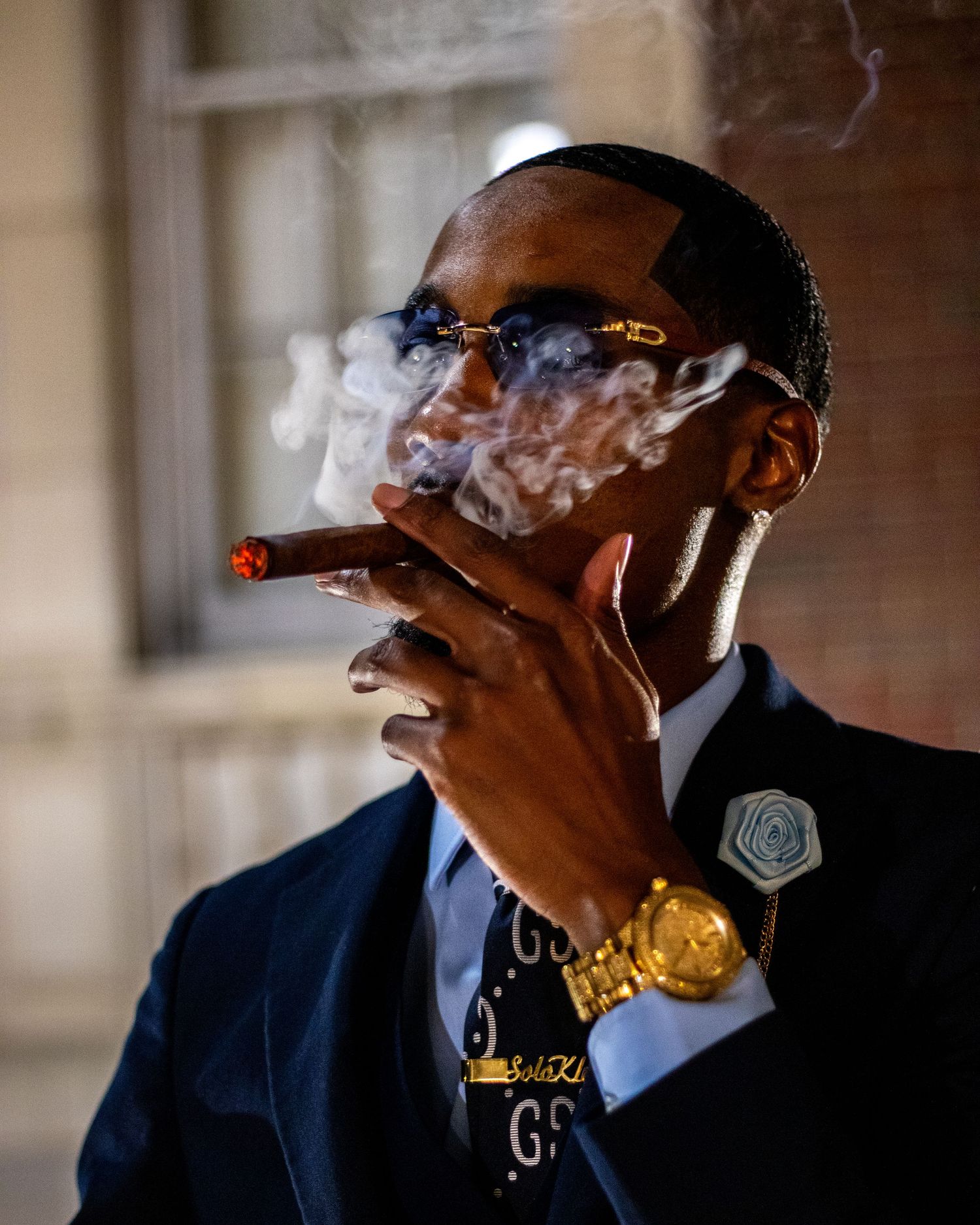 Well-dressed man smoking a cigar wearing gold watch and glasses in dramatic lighting.