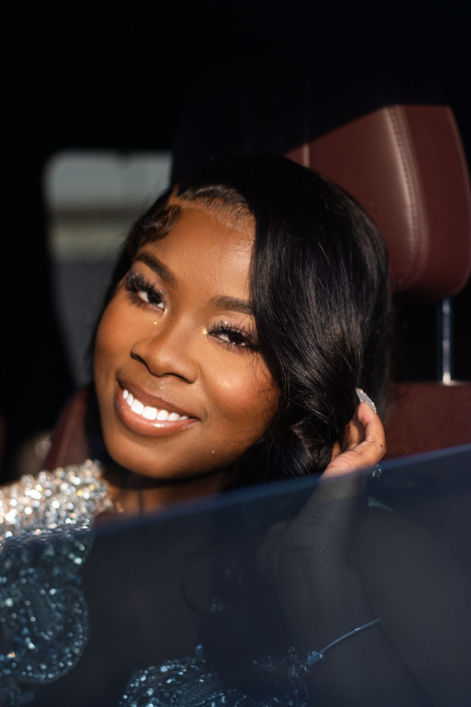 Smiling young woman with sleek black hair in a sparkling dress sits in a luxury car interior.