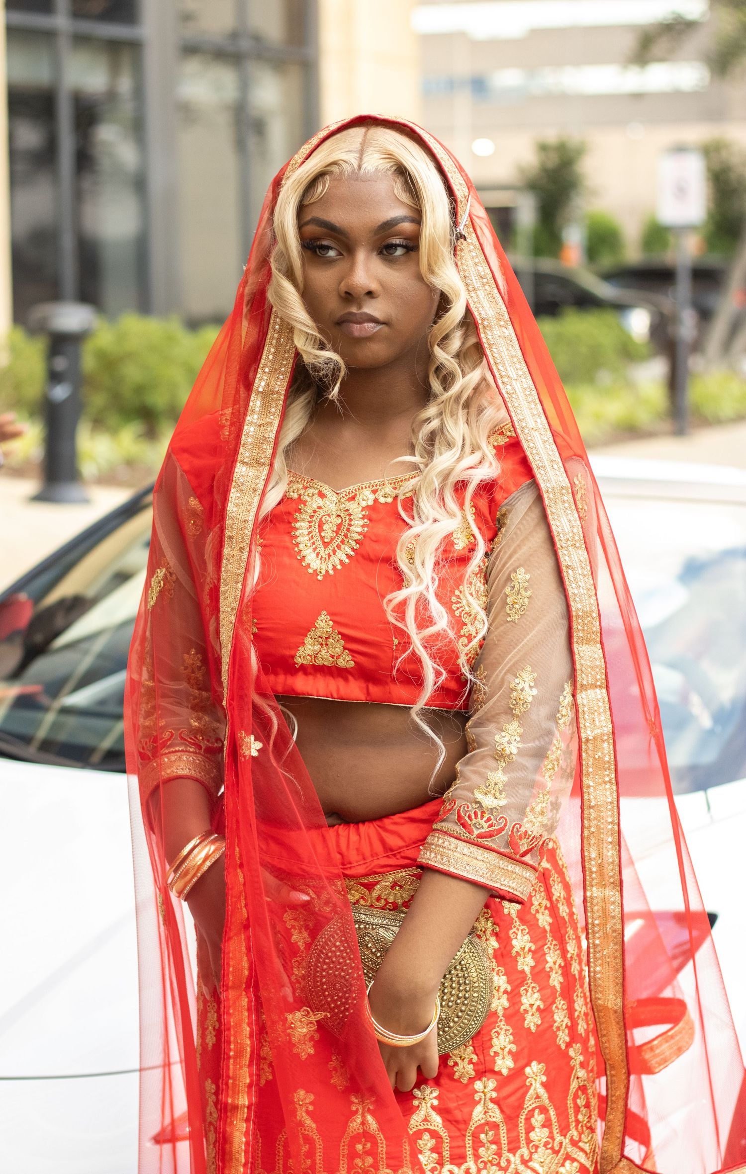 Young woman in ornate red and gold Indian lehenga with blonde hair and red veil poses outdoors.