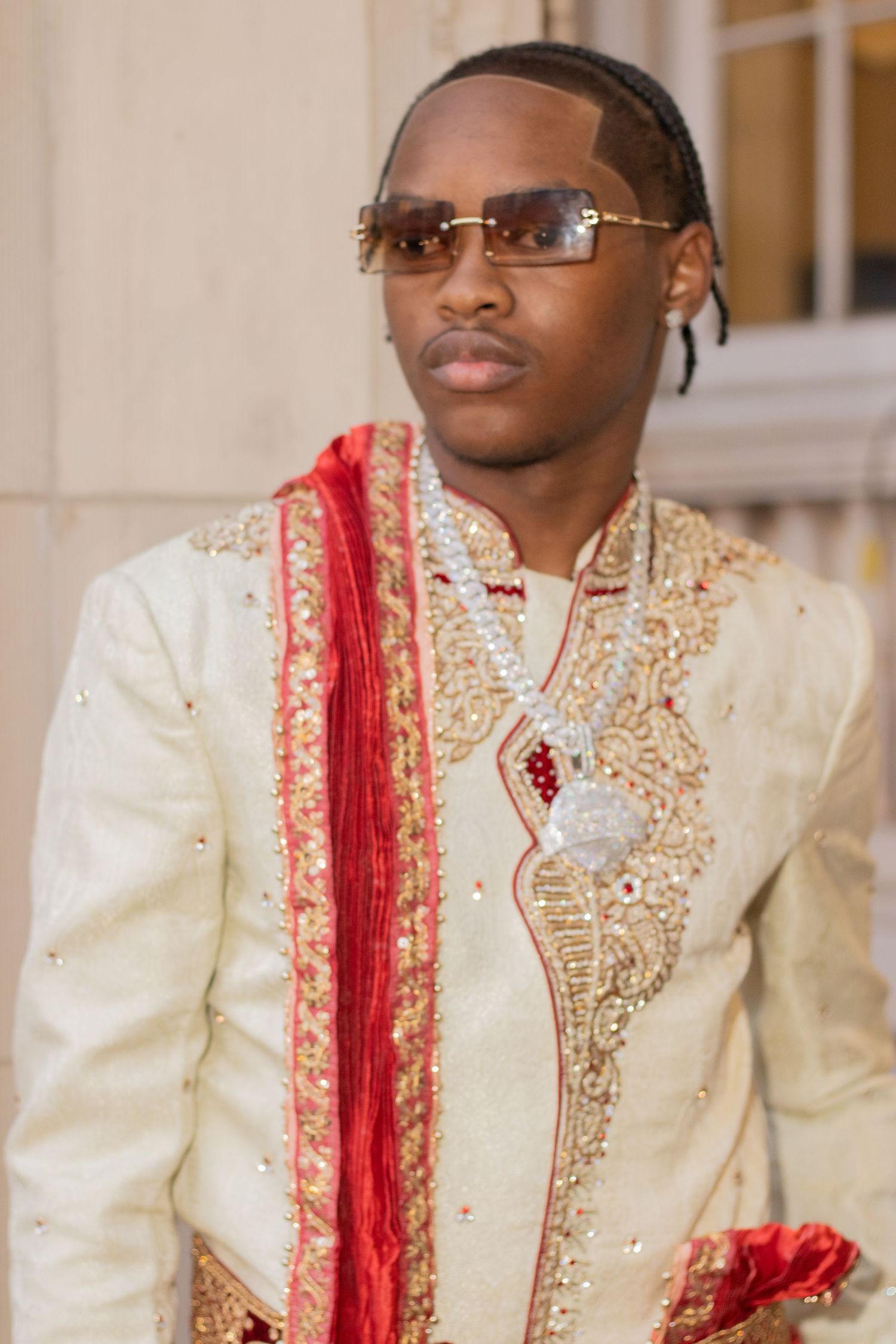Young man in cream and gold sherwani with red dupatta and sunglasses poses for a formal portrait.