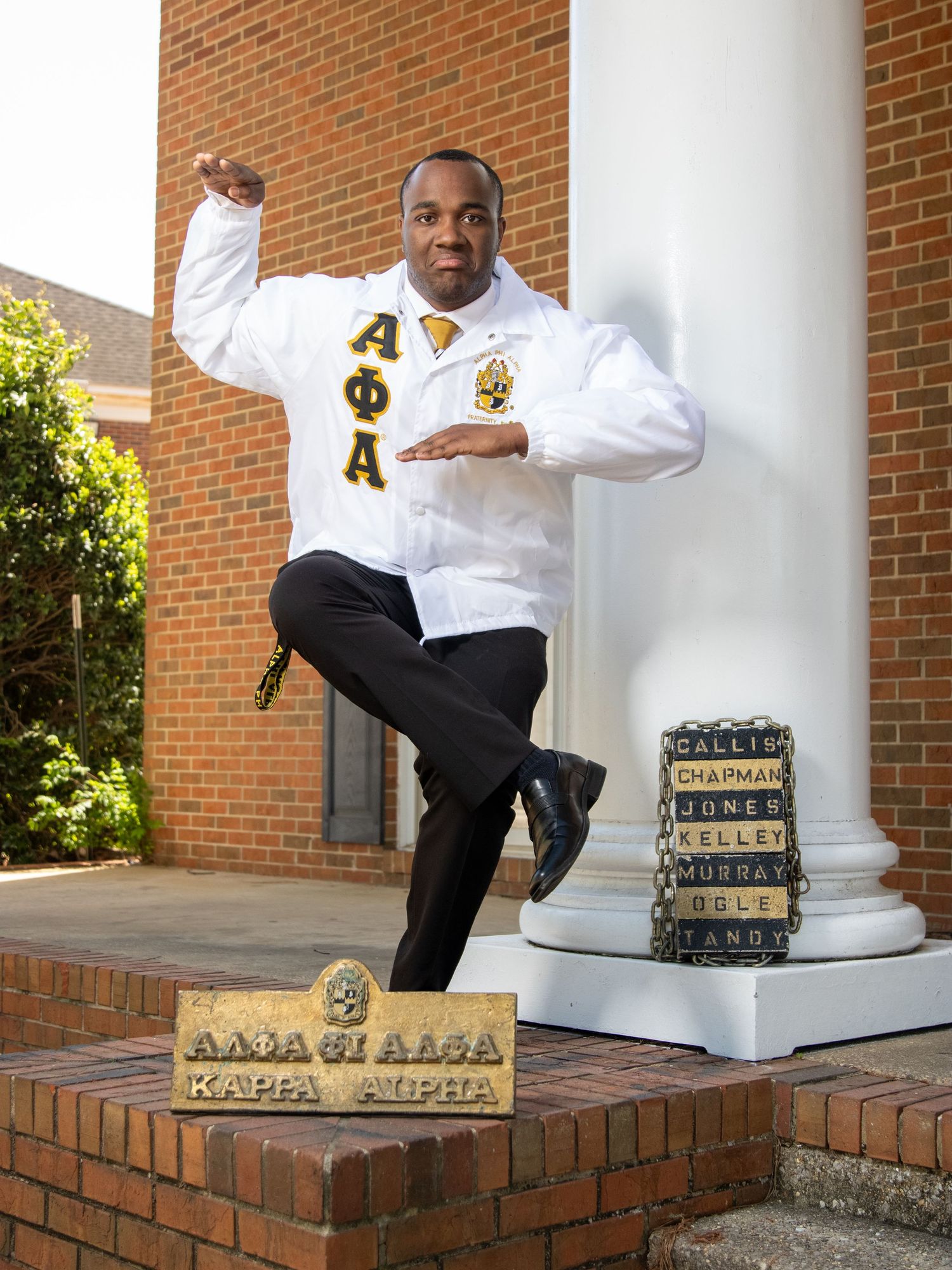 Fraternity member in white jacket and black pants poses energetically on brick steps with organization blocks.