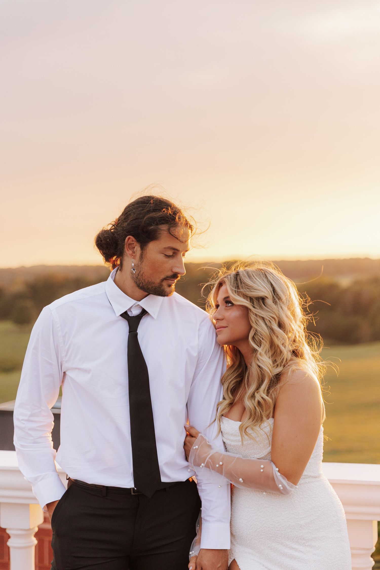 A couple in formal wedding attire embrace on a balcony against a warm sunset sky.
