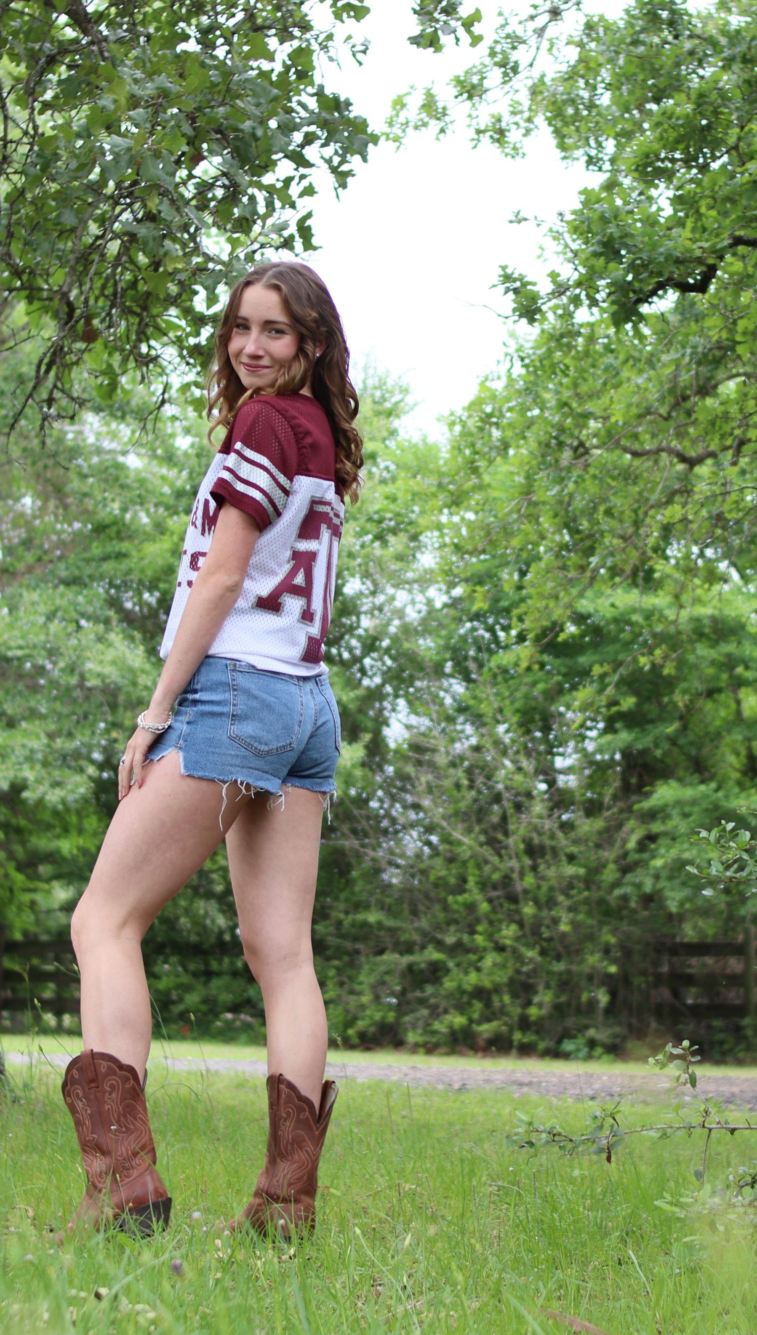 Young woman in cowboy boots, denim shorts, and maroon jersey poses outdoors near green trees.