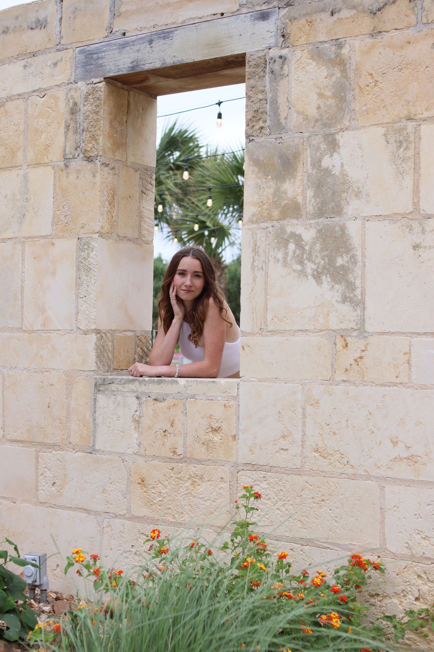 A young woman leans thoughtfully through a rustic stone wall window framed by string lights and orange flowers.