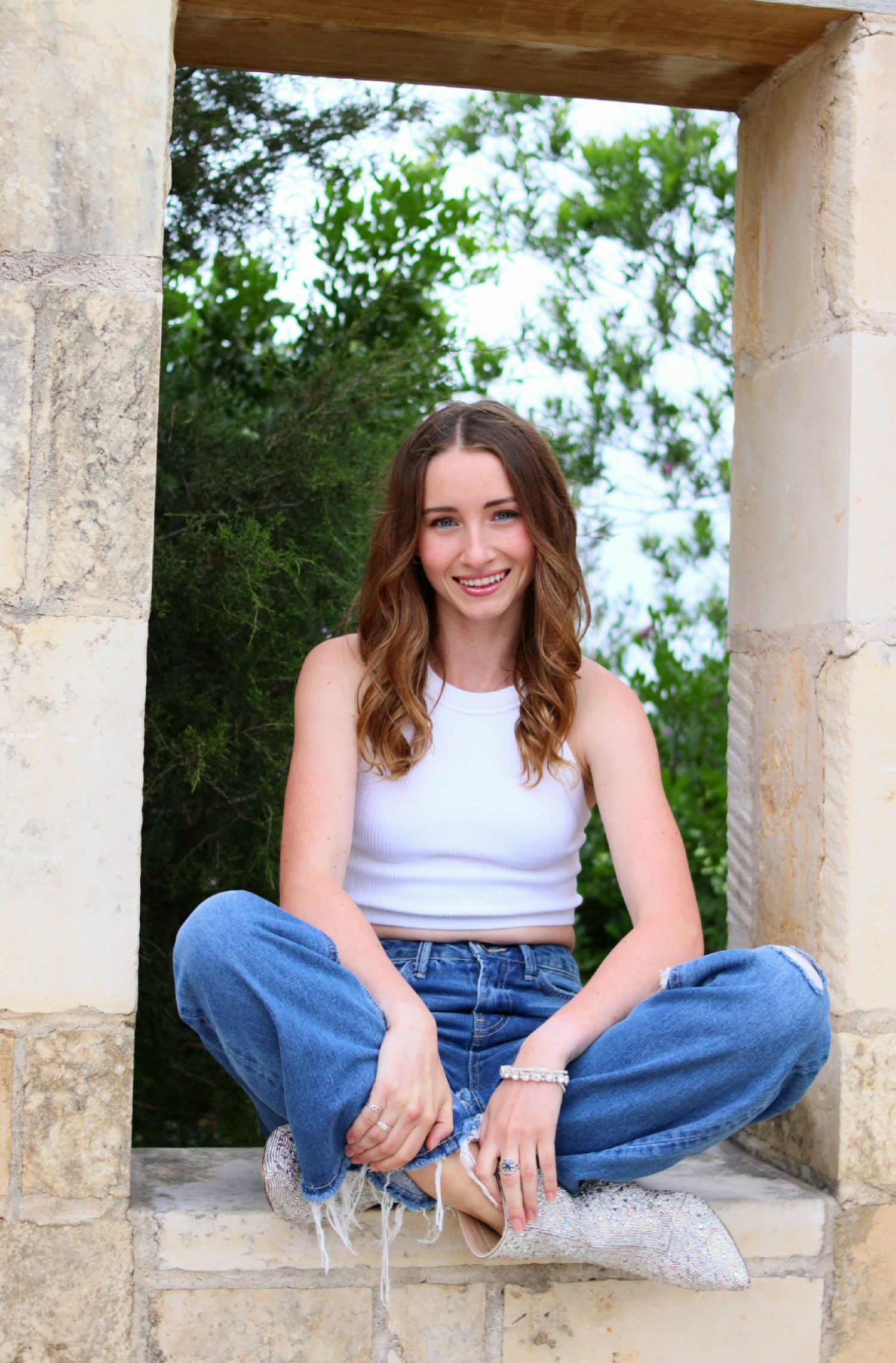 A smiling young woman in a white tank top and jeans sits cross-legged in a stone archway outdoors.
