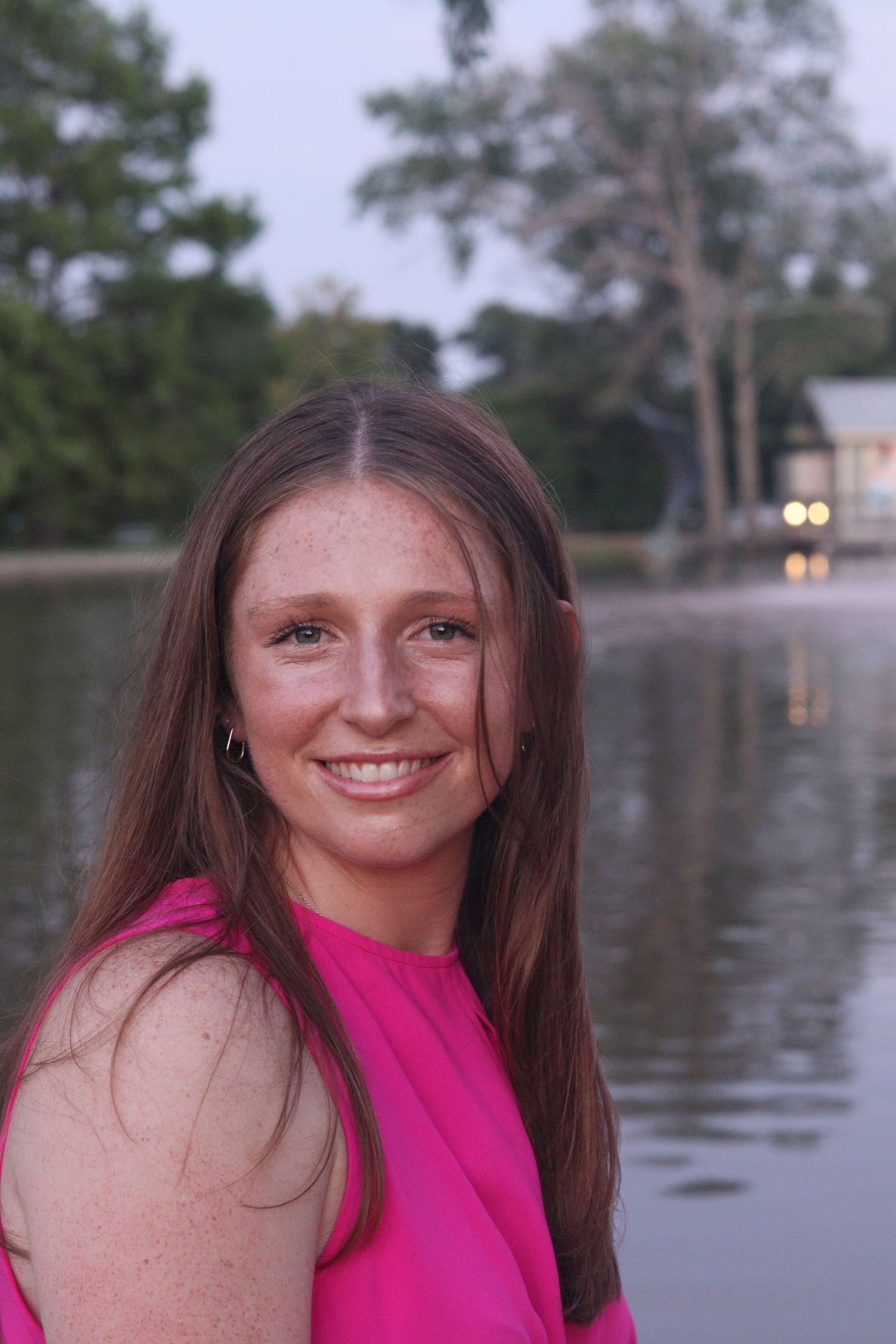 A smiling young woman with freckles in a pink dress poses near a reflective lake at dusk.
