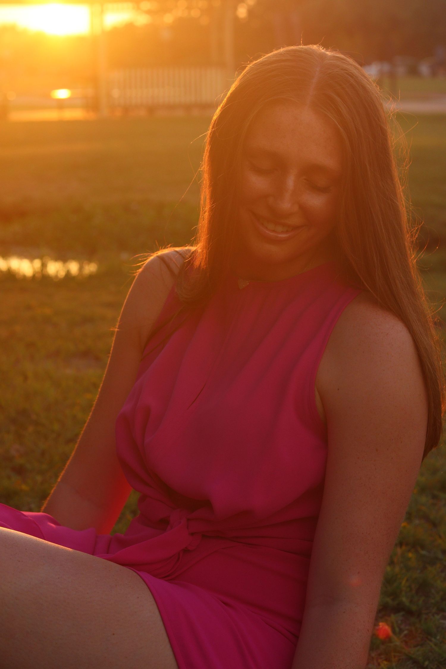 A young woman in a pink dress smiles while sitting on grass bathed in warm golden sunset light.
