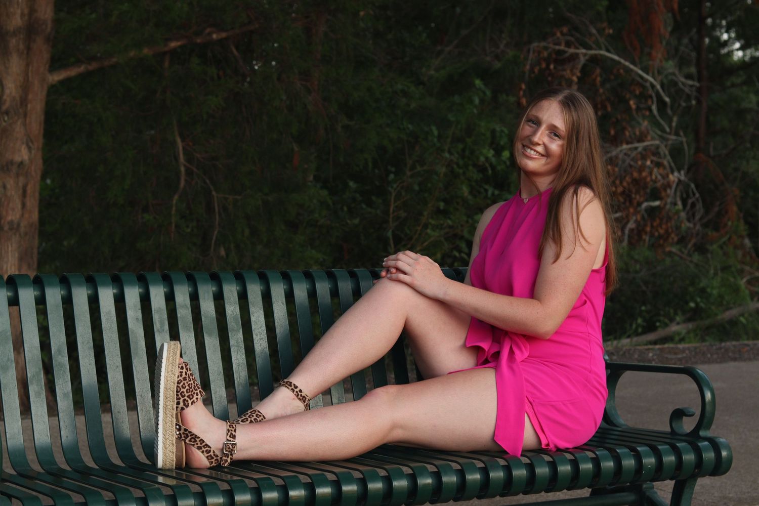 A smiling young woman in a pink mini dress sits relaxed on a green park bench wearing leopard sandals.
