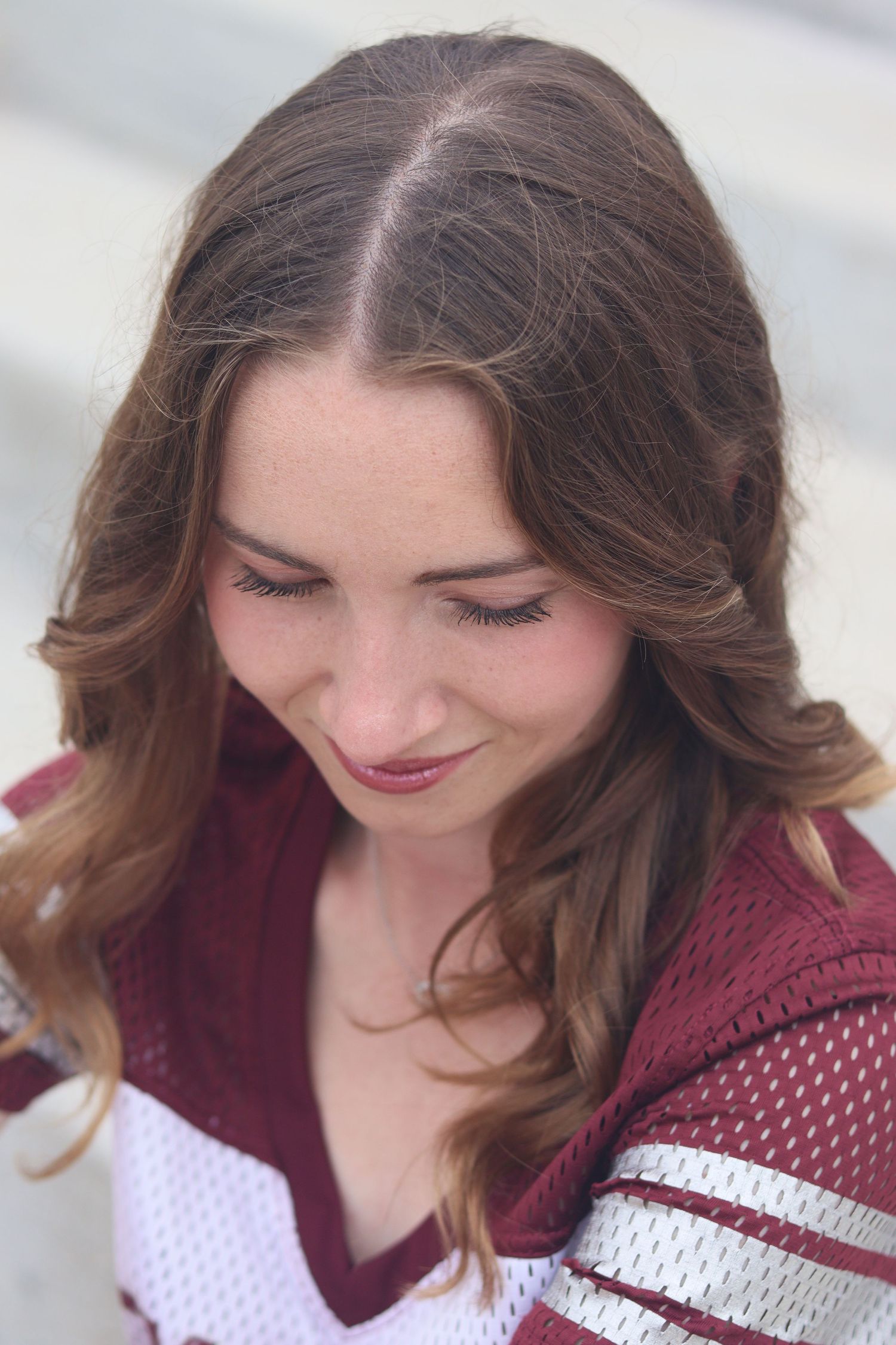 Young woman with wavy brown hair looking down wearing a maroon and white varsity jersey.