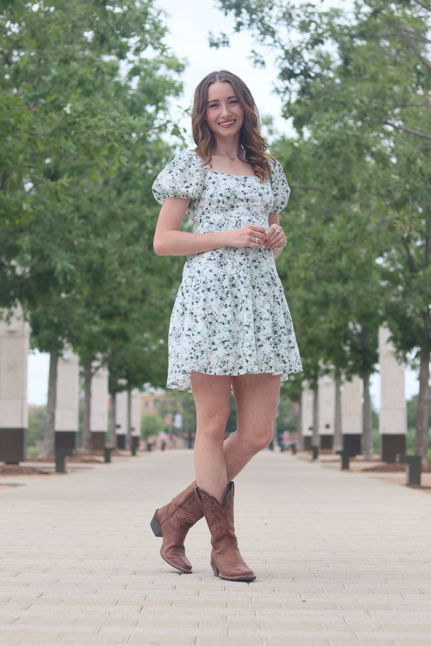 Young woman in floral mini dress and brown cowboy boots smiling on a tree-lined walkway.