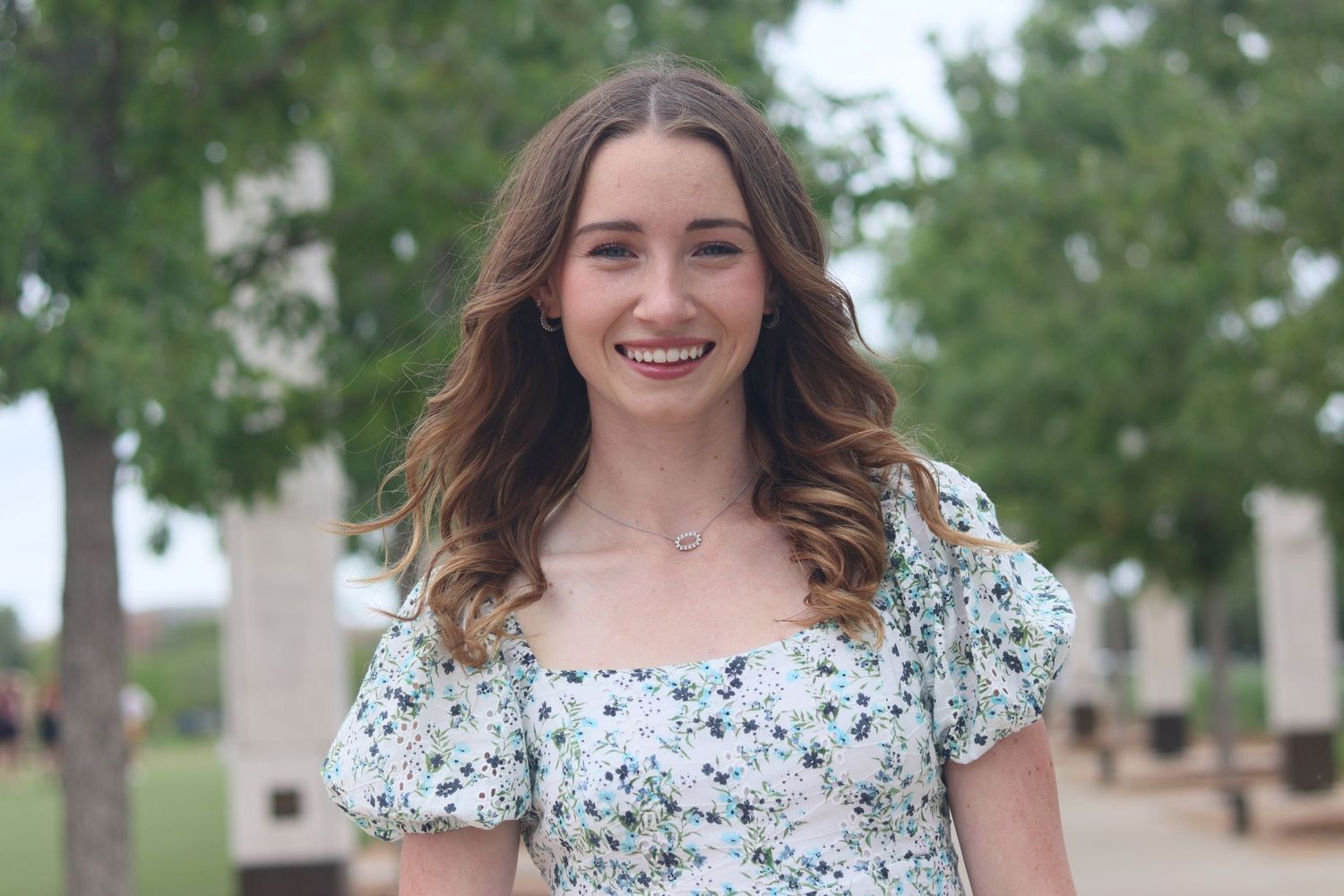 Smiling young woman in floral dress with wavy brown hair posing outdoors among green trees.