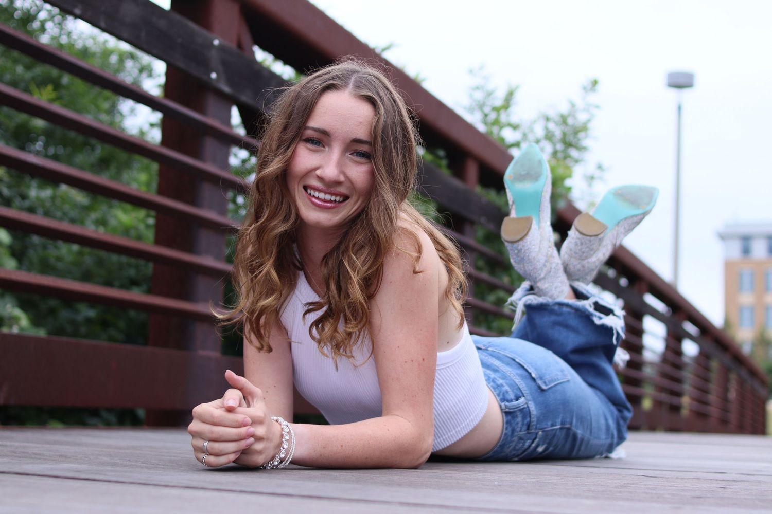 Smiling young woman lying on a wooden bridge deck in jeans and white top with colorful sneakers.