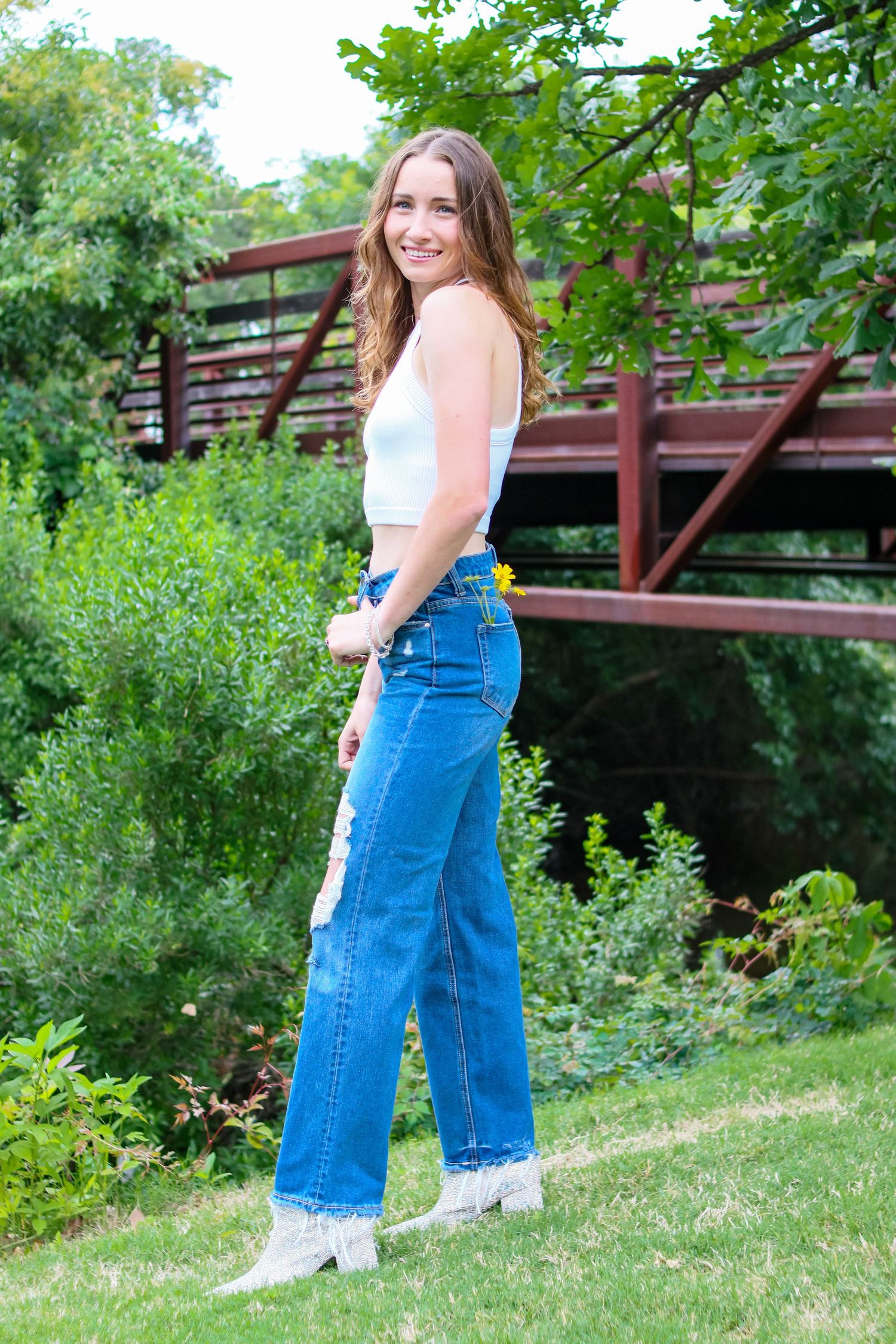 Young woman in white crop top and blue jeans smiling near a red bridge in a garden.