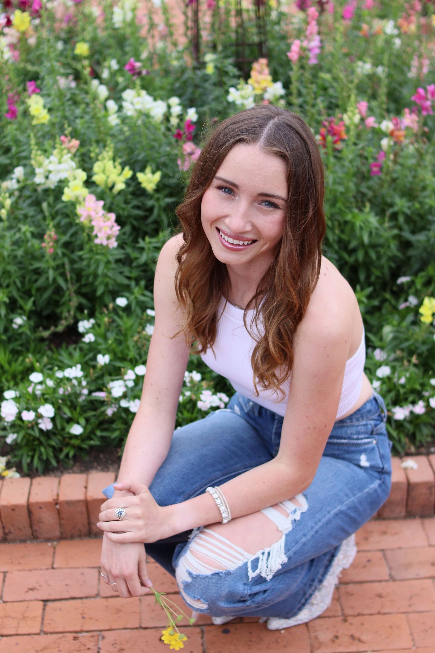Smiling young woman in white top and ripped jeans crouching in front of colorful flowers.