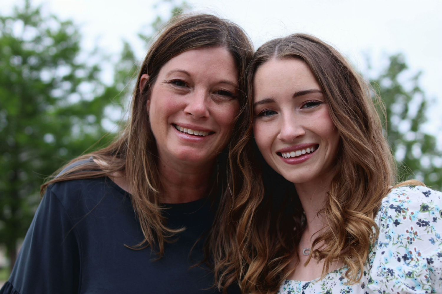 Two smiling women posing together outdoors with green trees in the background.