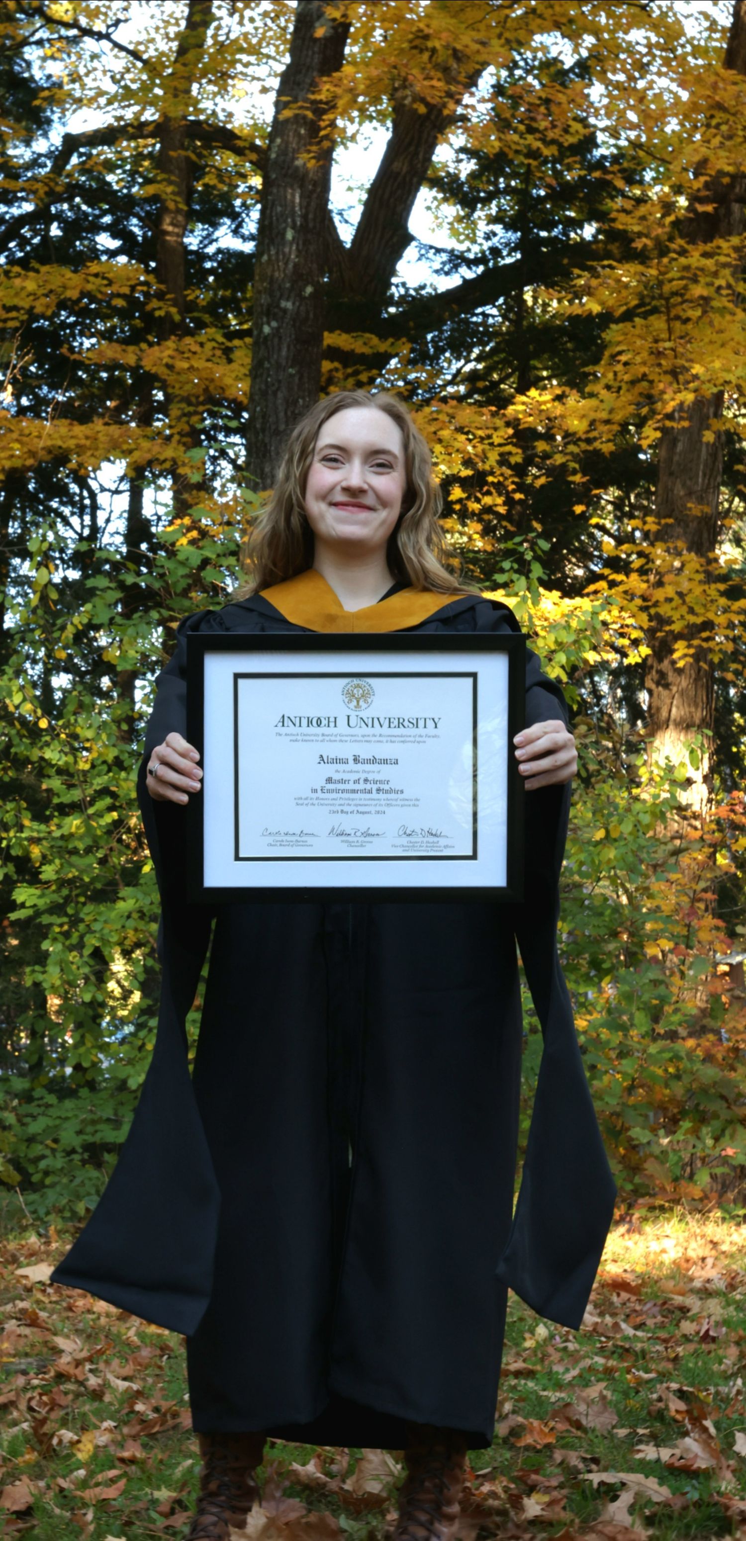 Proud graduate in black cap and gown holding a framed diploma outdoors in autumn.