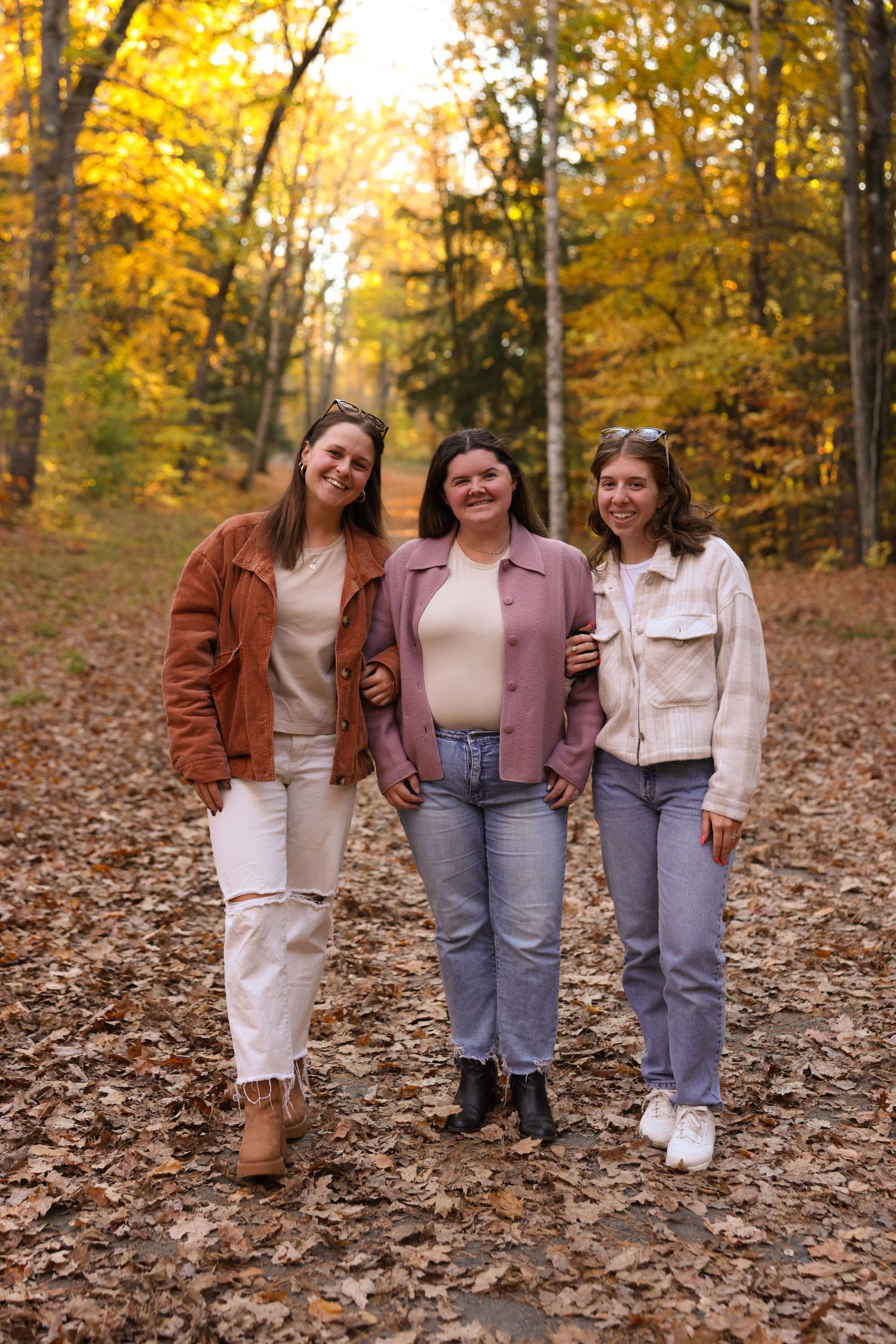 Three smiling young women walking together on a leaf-covered path in an autumn forest.