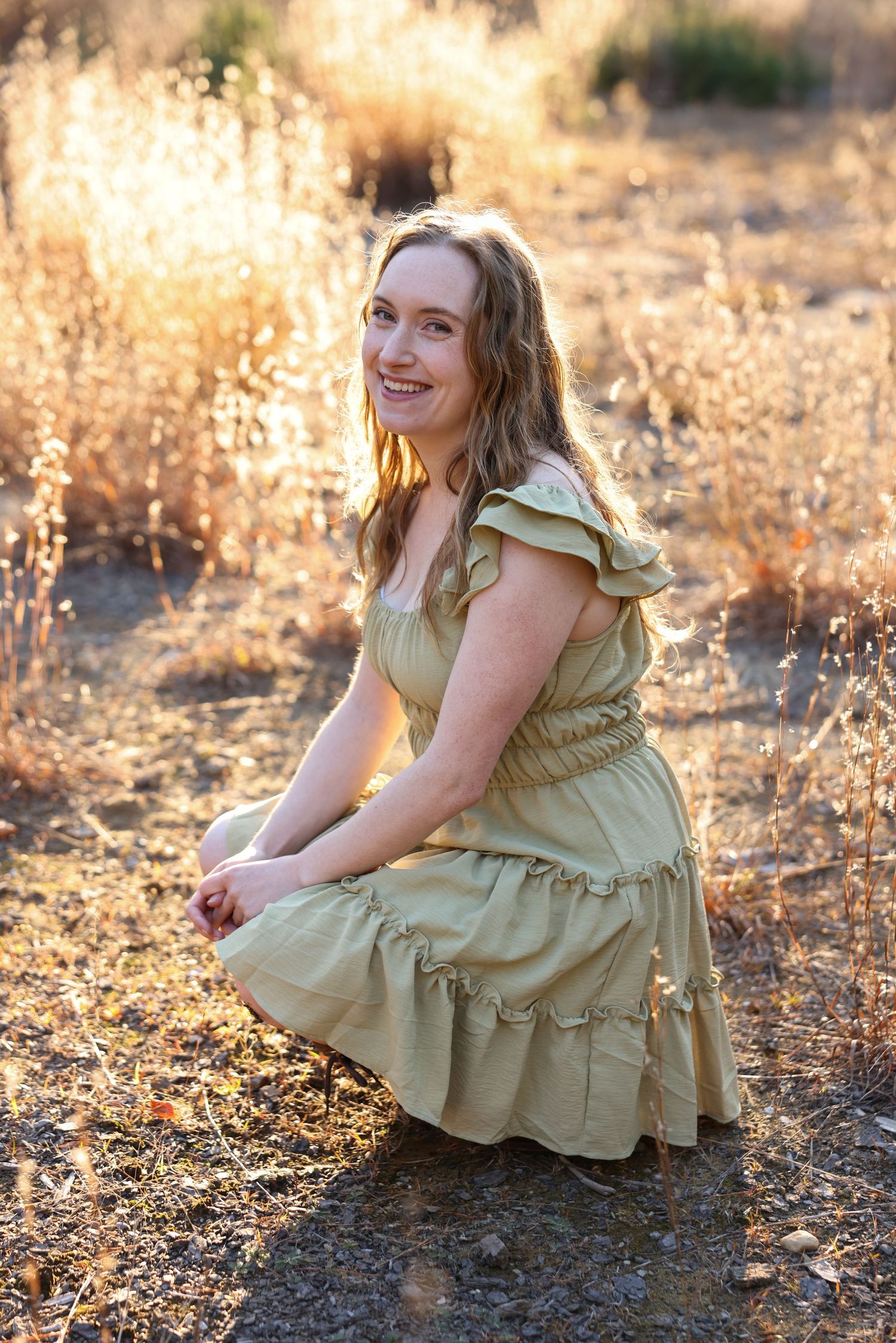 Smiling woman in a sage green ruffled dress crouching in a golden sunlit field.