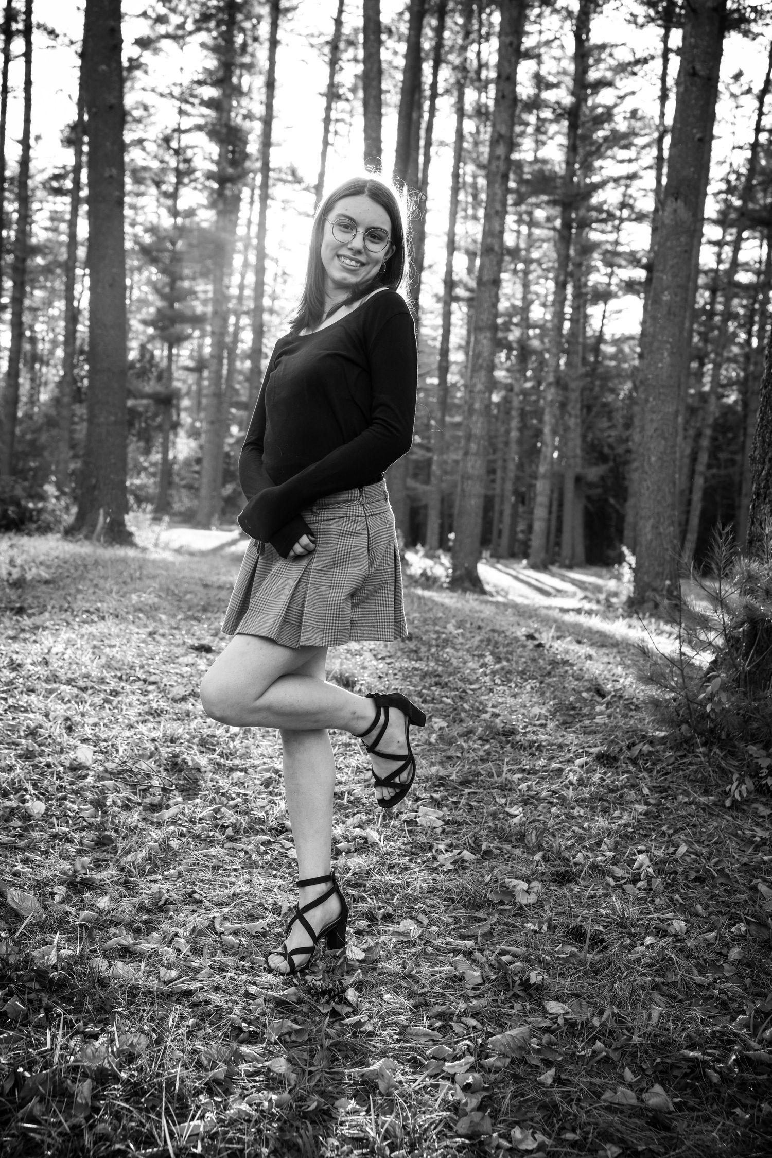 Young woman in glasses, black top and plaid skirt posing in forest in black and white photo.