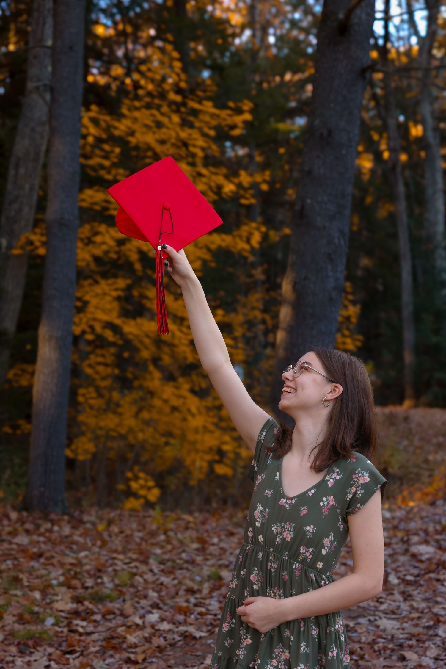 Smiling graduate tossing red mortarboard cap in autumn forest with colorful fall foliage.