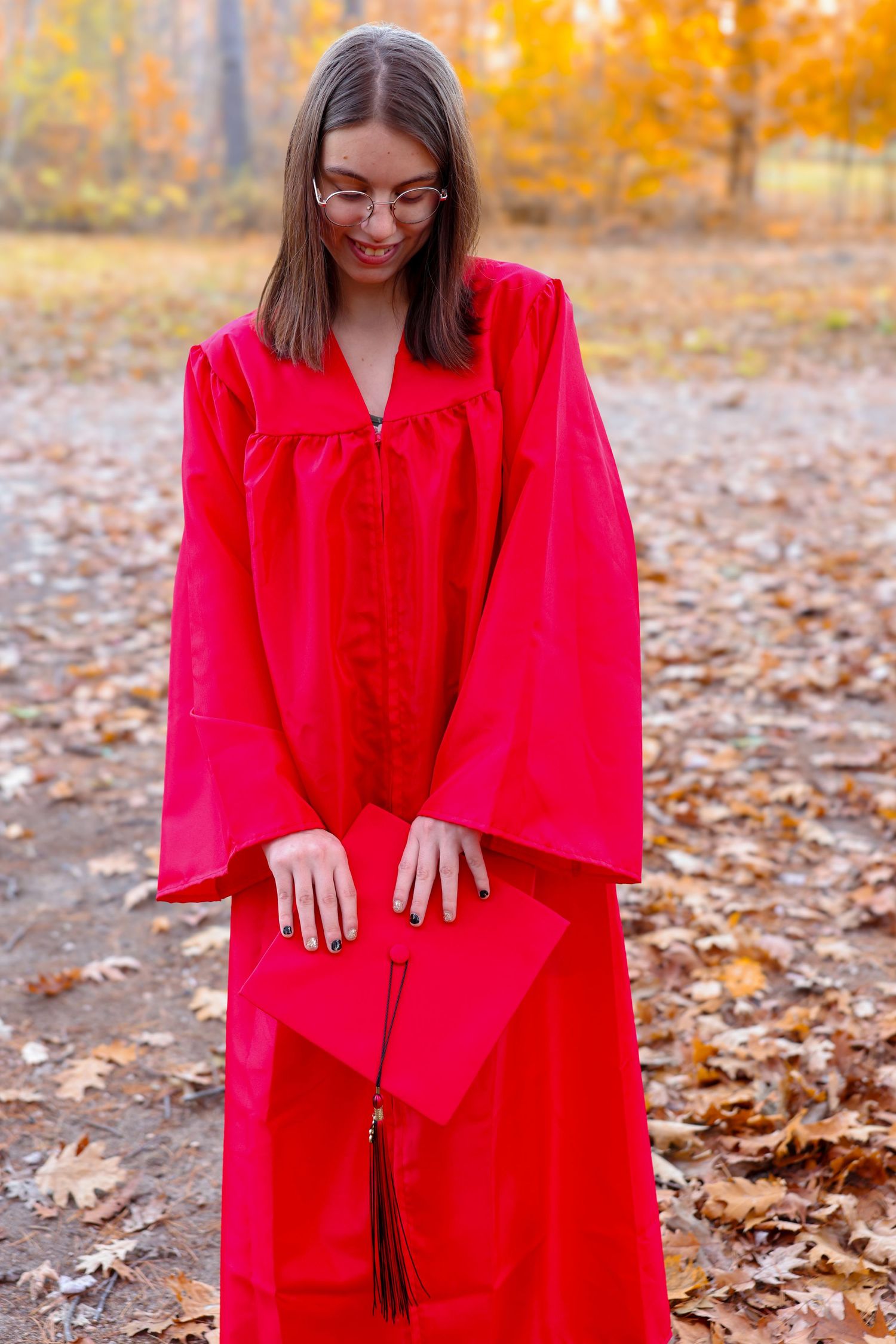 Young woman in red graduation gown and cap standing among fallen leaves in autumn forest.