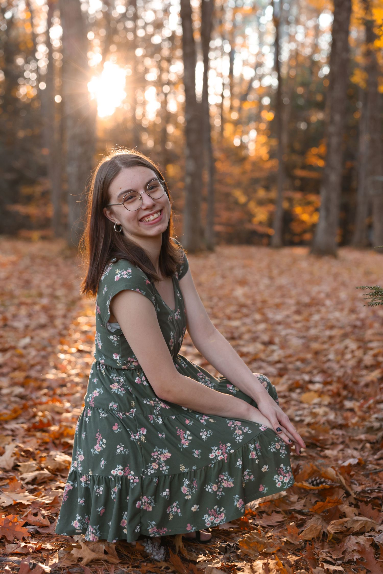Smiling brunette woman in floral green dress crouching among fall leaves in golden sunset forest.
