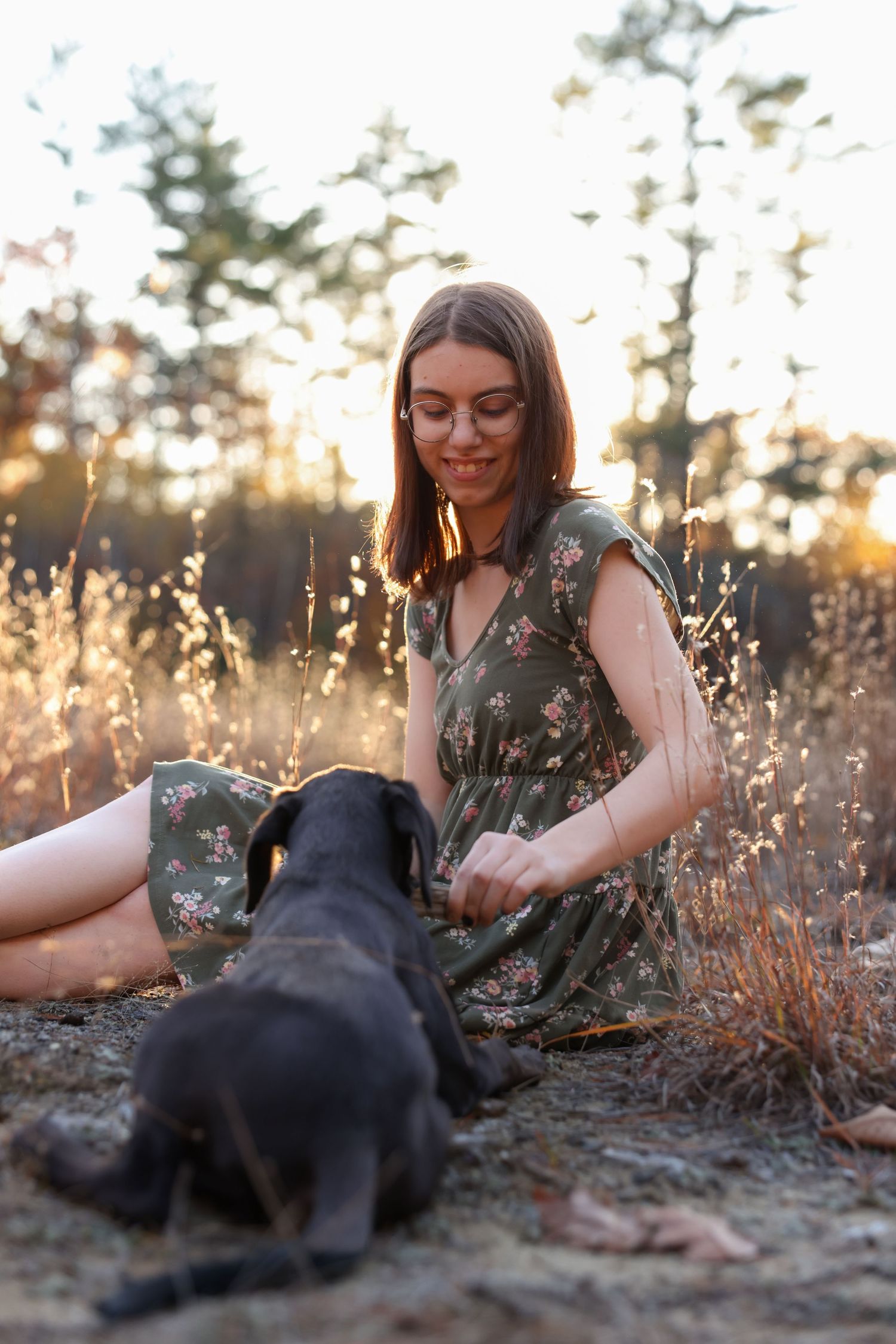 Young woman in floral green dress sitting outdoors at sunset playing with a black puppy.