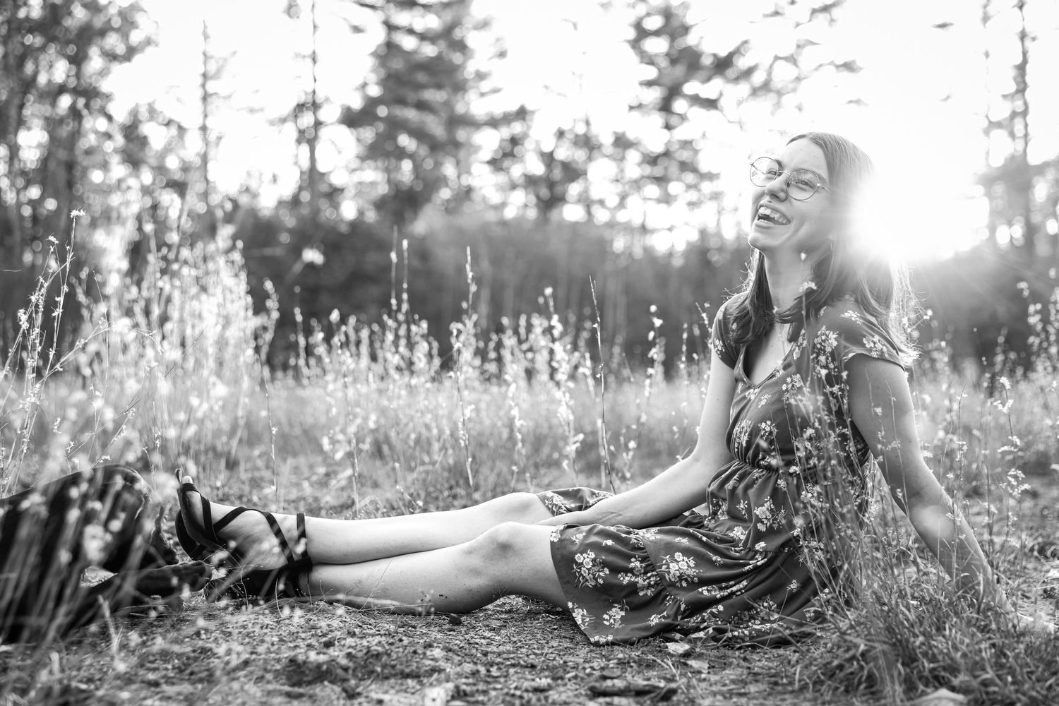 Black and white photo of a smiling woman in a floral dress sitting in a sunlit meadow.