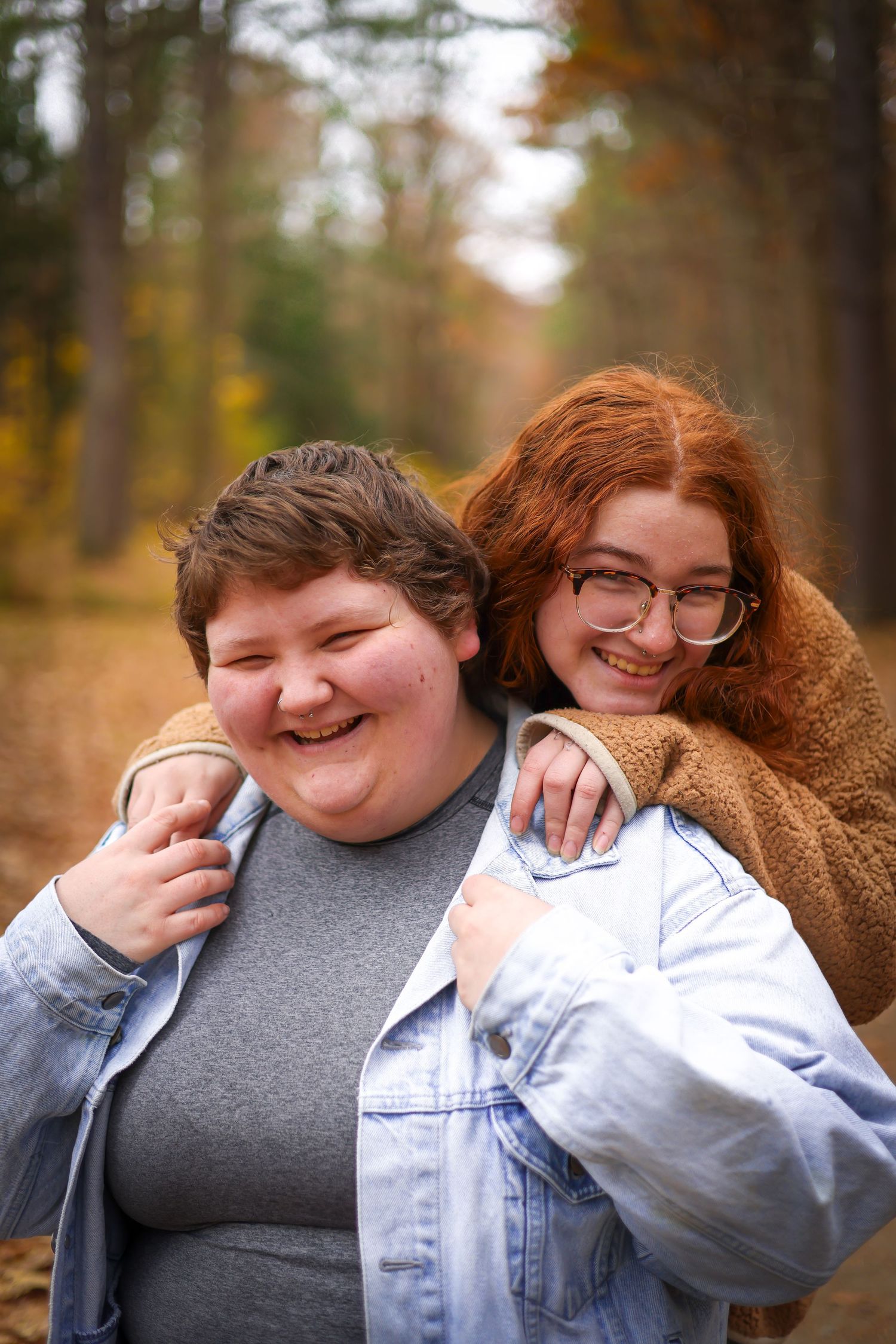 Two smiling teenagers pose together in an autumn forest, one hugging the other from behind.