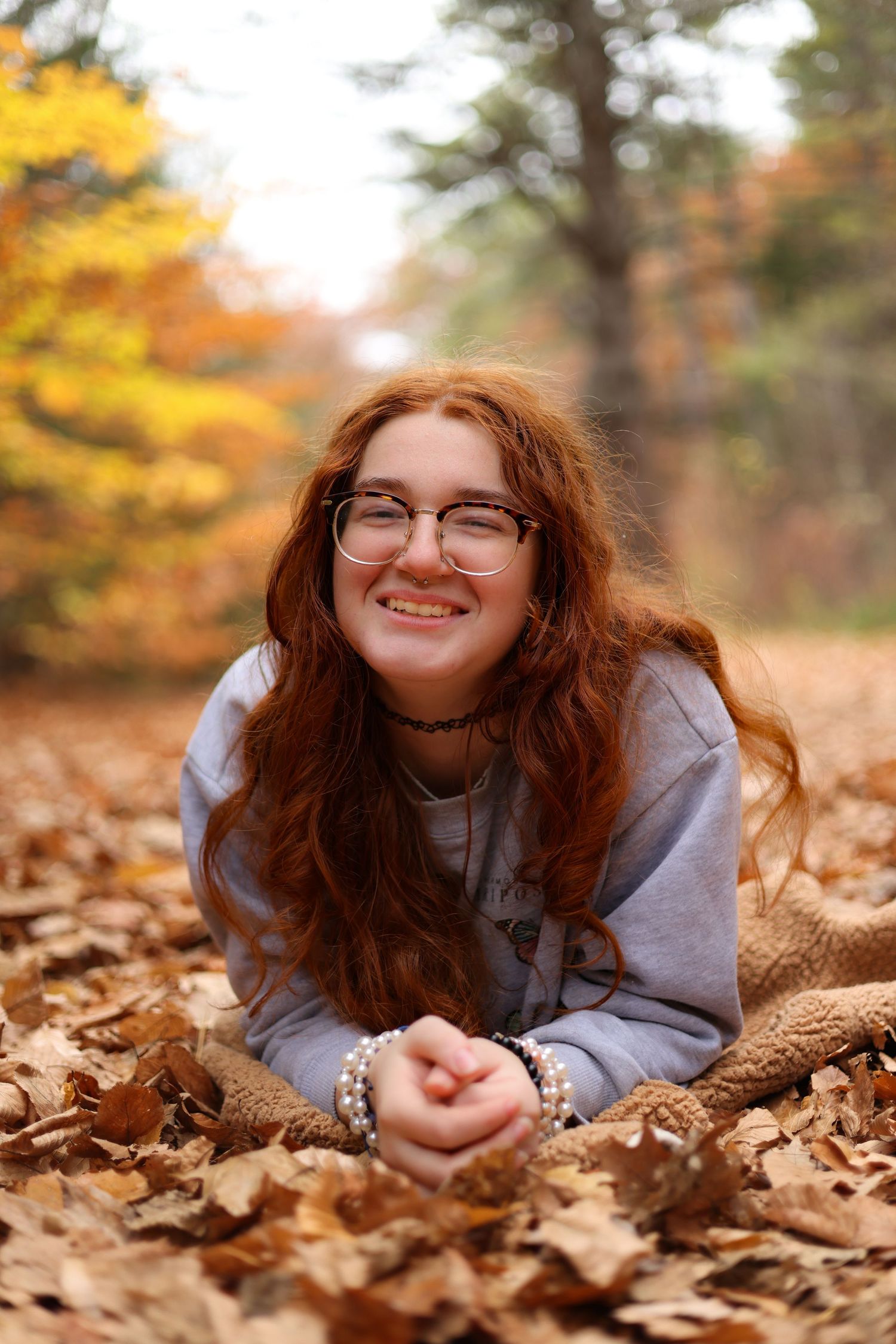 A smiling redhead teenage girl with glasses lies in autumn leaves in a colorful fall forest.