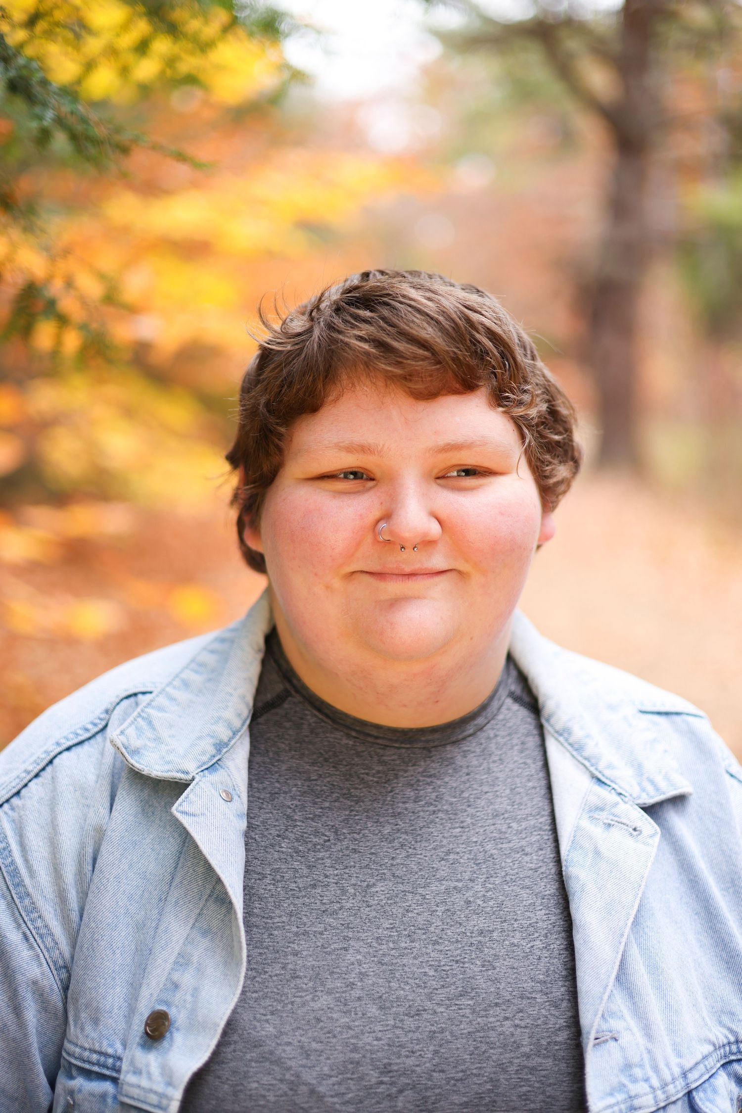 A young person with short brown hair smiles softly outdoors among vibrant autumn foliage.