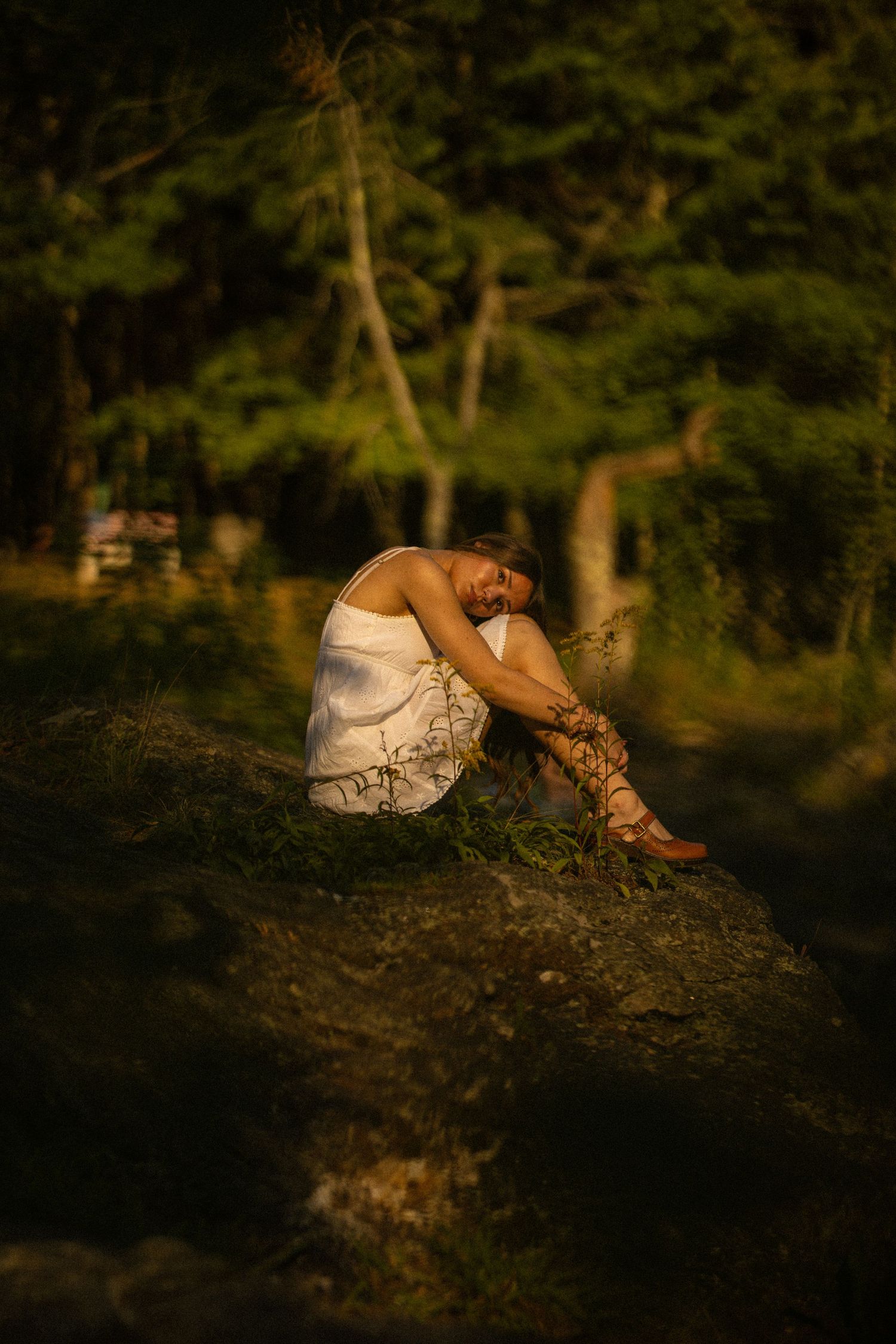A person in a white dress and gladiator sandals sits curled up on a rock in a sunlit forest setting.