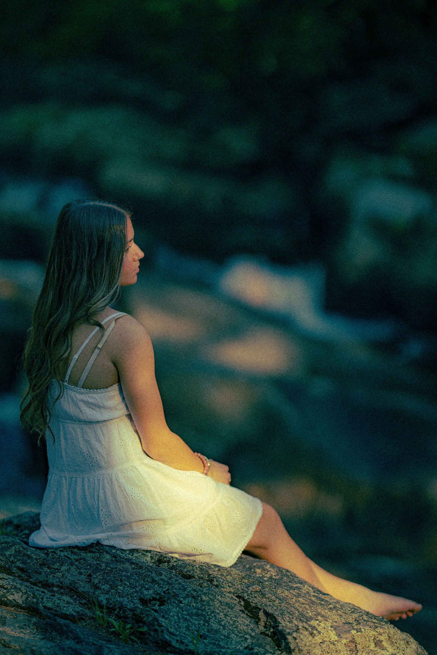 A barefoot young woman in a white dress sits peacefully on rocks beside a flowing stream at dusk.