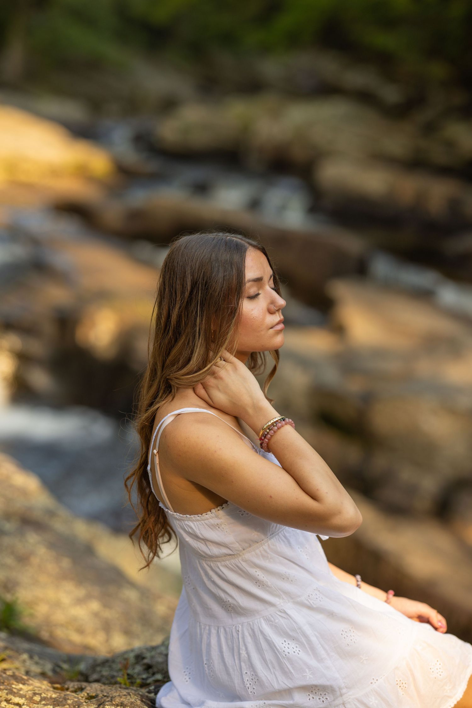 Young woman in white dress sitting peacefully by a rocky waterfall with eyes closed, wearing bracelets.