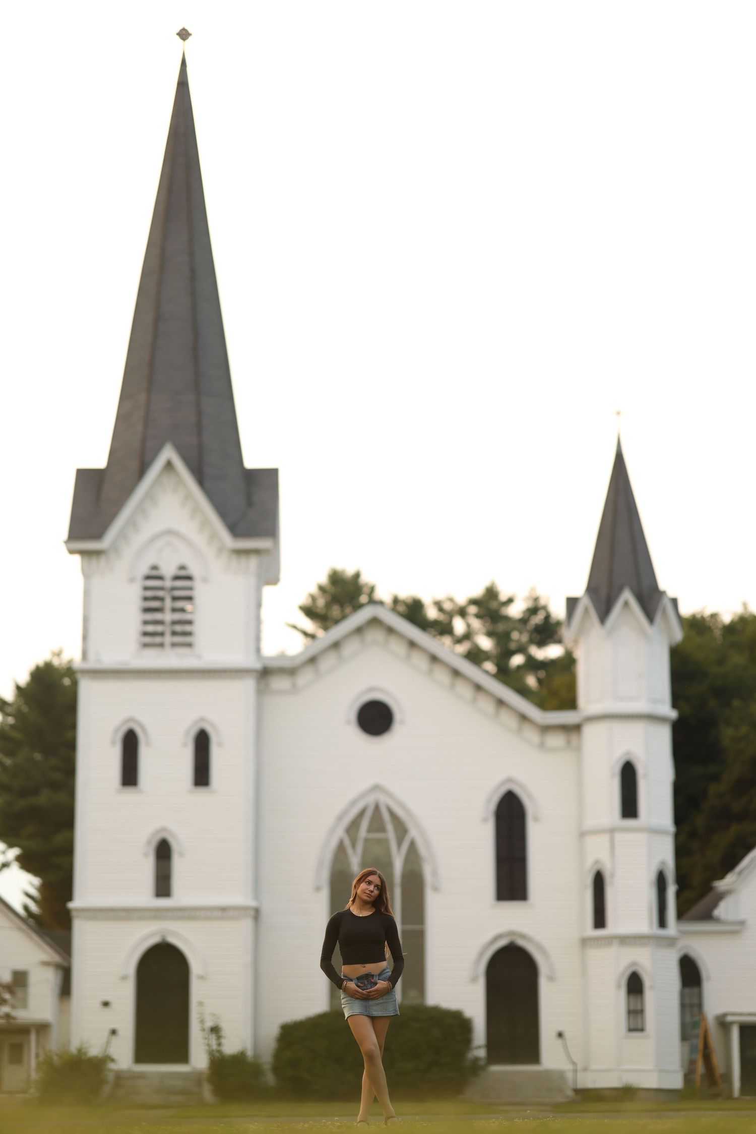 Young woman in black top and denim skirt standing in front of a white historic church with tall steeples.