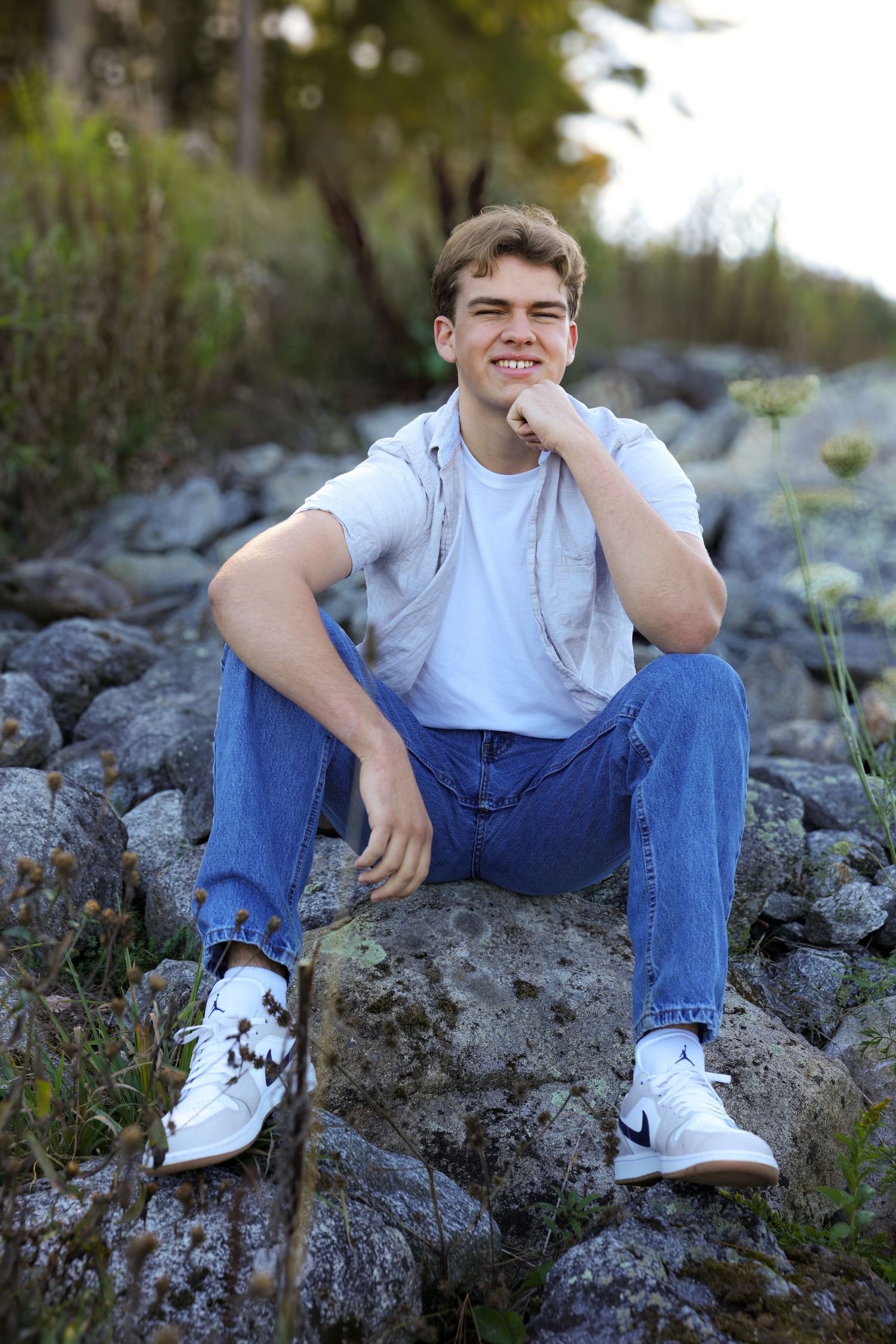 Smiling young man in white tee and jeans sits on rocks outdoors in natural wooded setting.