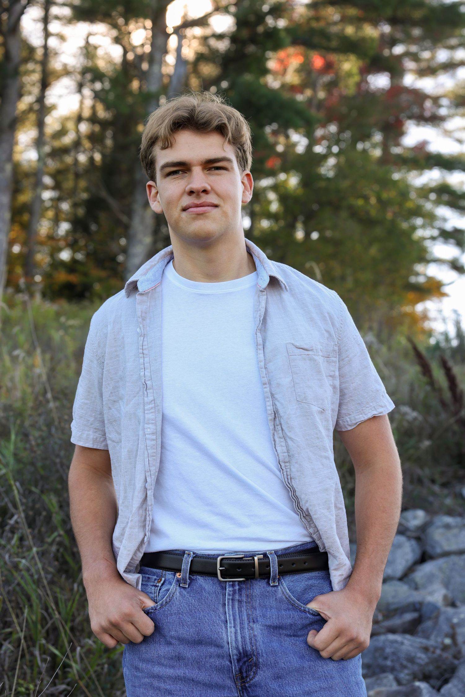 Young man in open linen shirt over white tee and jeans stands outdoors near rocky wooded area.