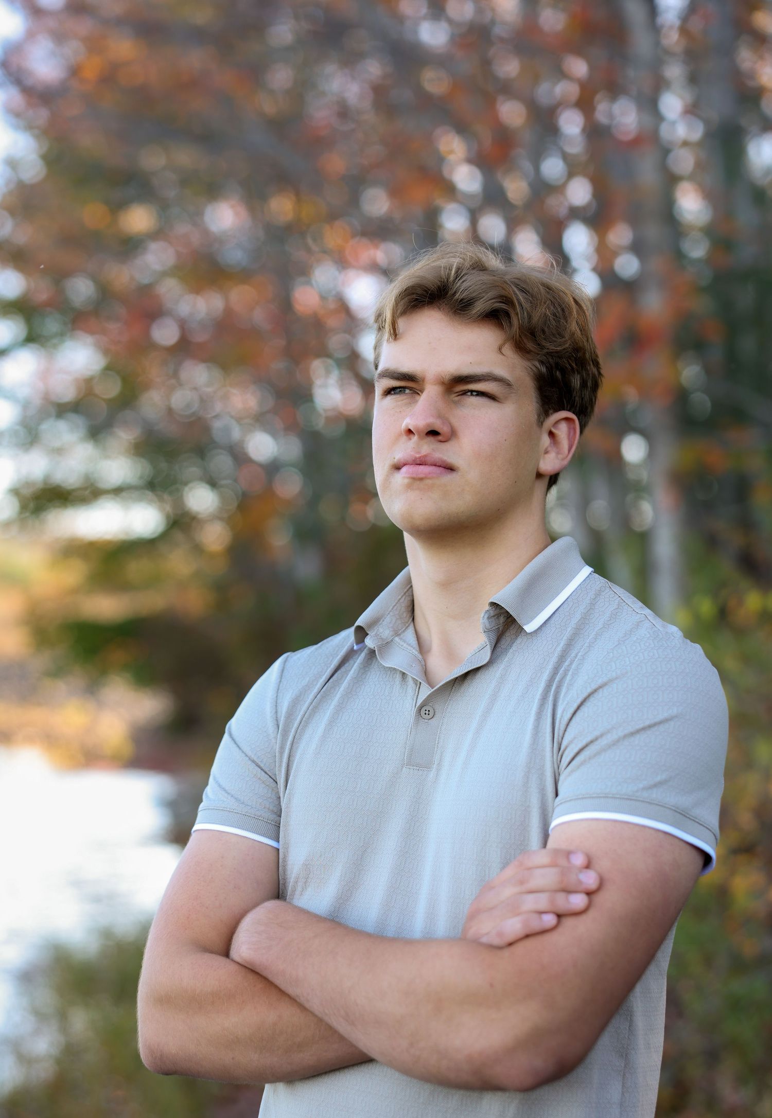 Young man in gray polo with arms crossed stands confidently outdoors with colorful autumn trees behind.