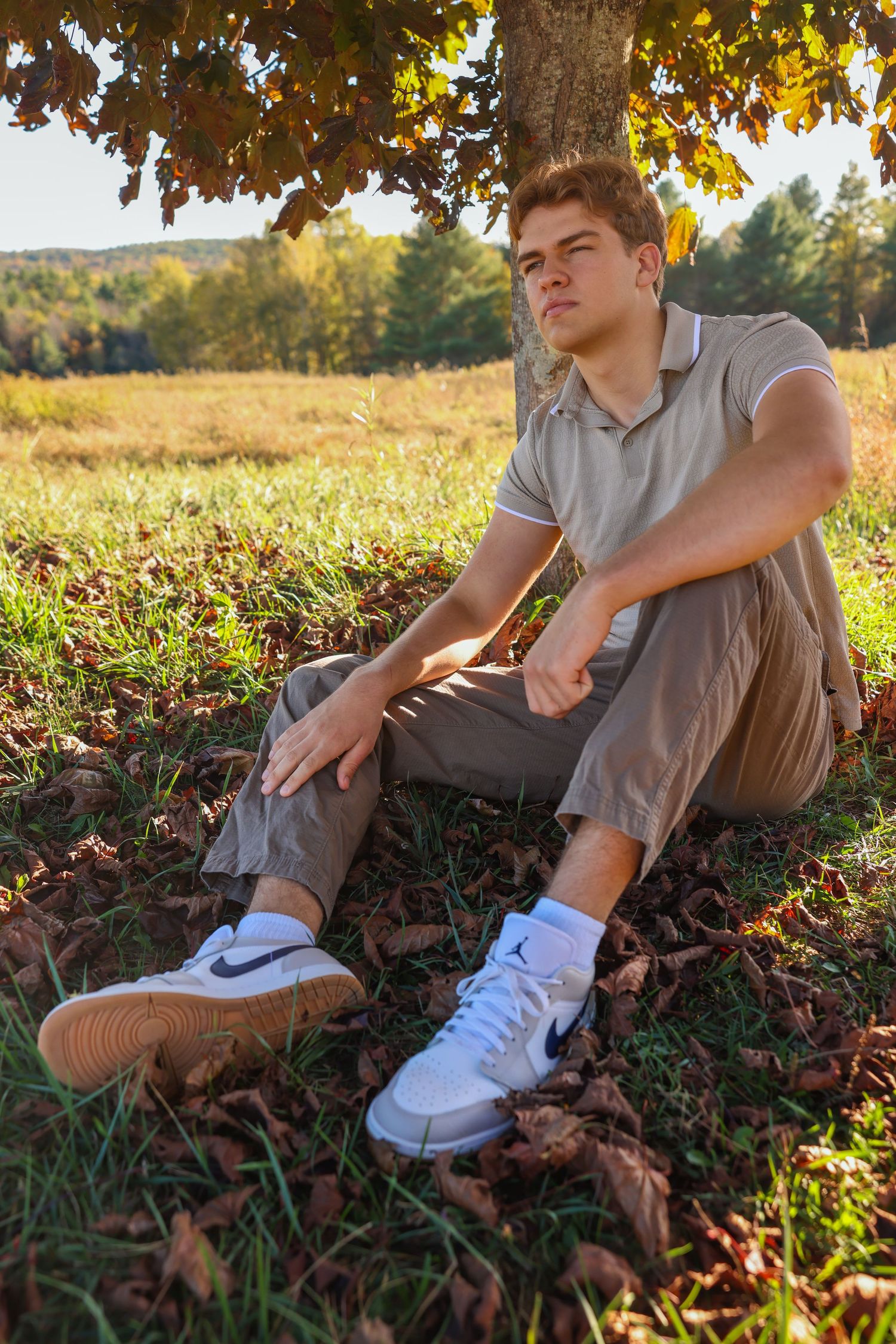 Young man in beige polo and khakis sits under tree in golden autumn field with fallen leaves.