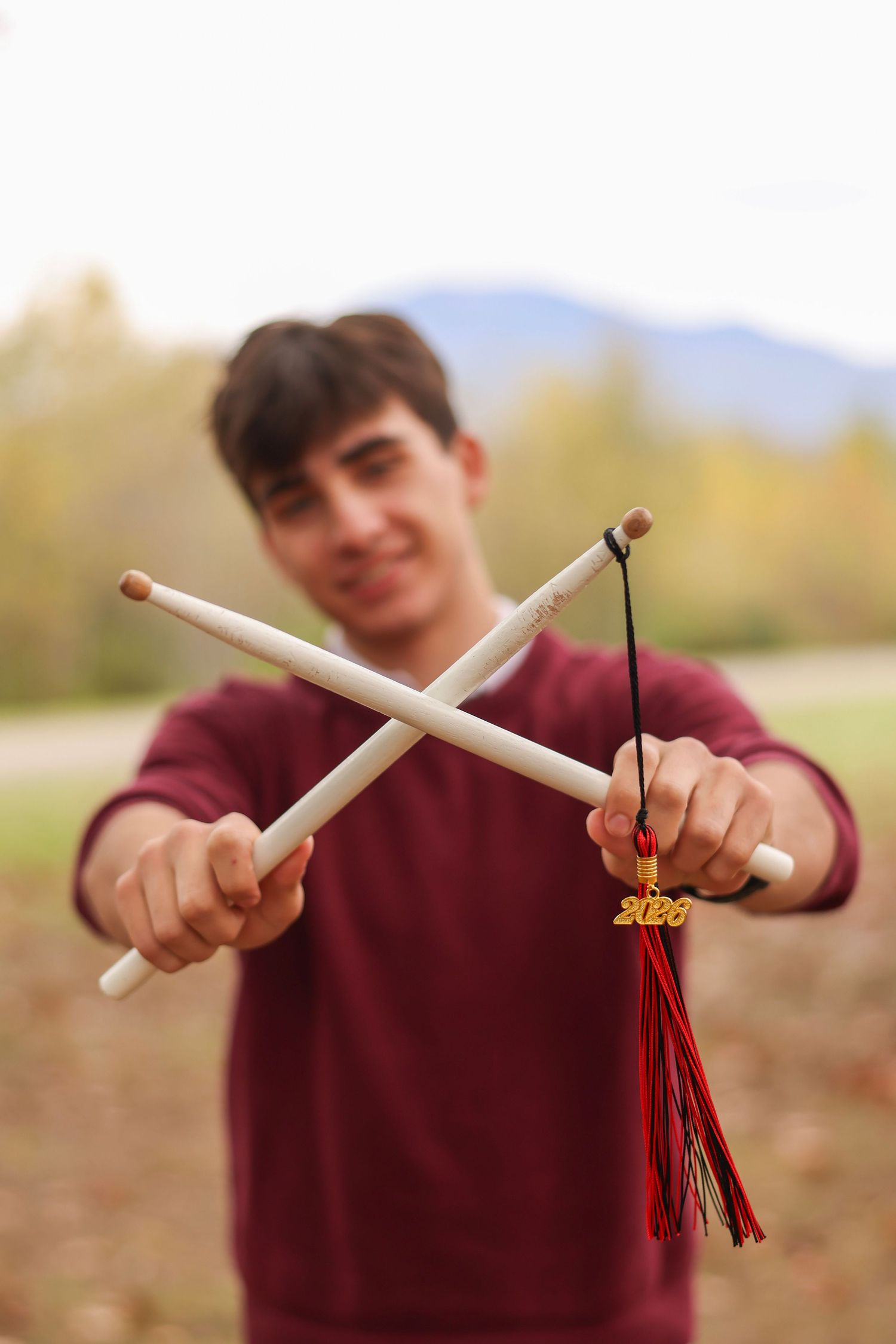 Teen boy holding crossed drumsticks with a 2020 graduation tassel in an outdoor setting.