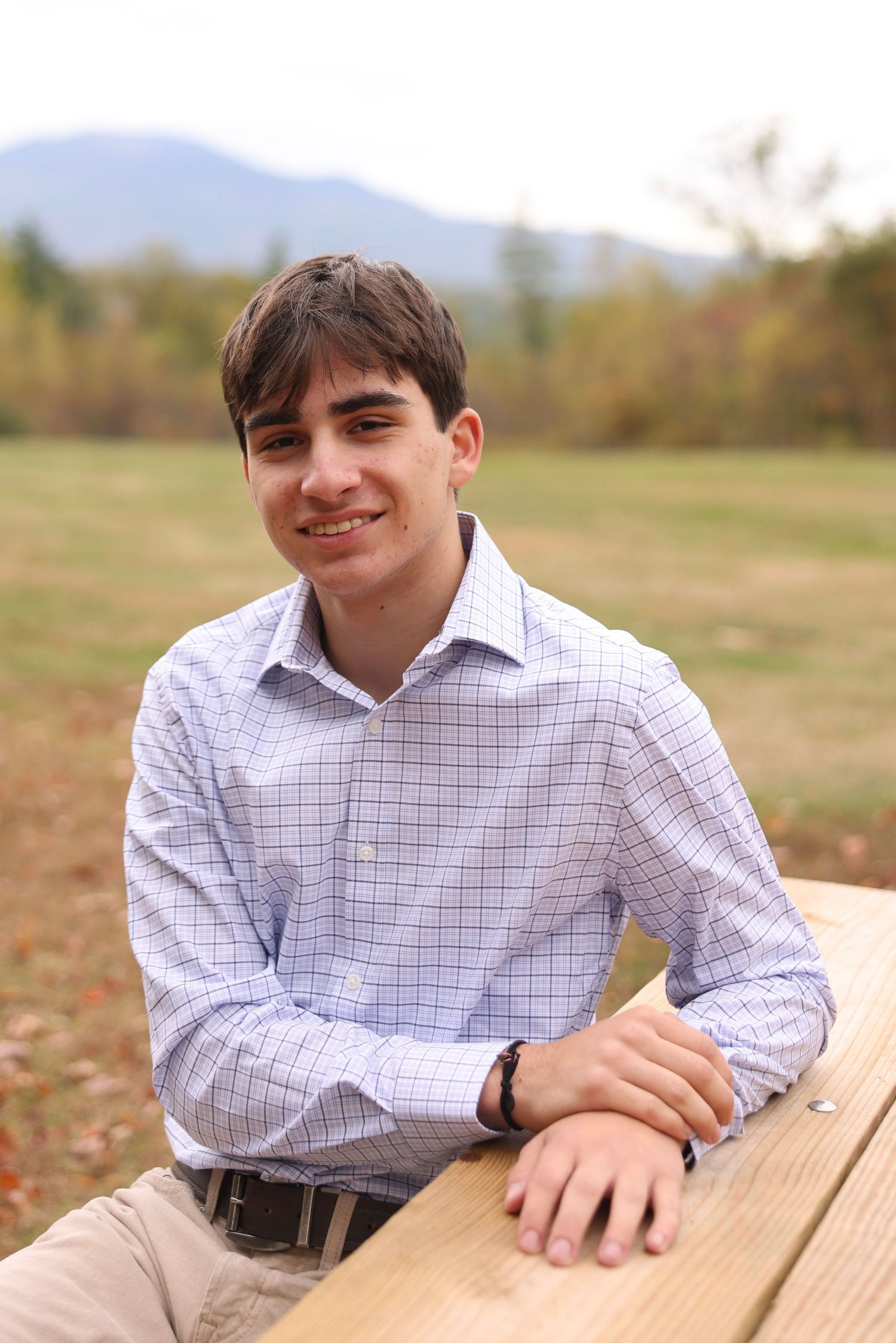 Smiling teenage boy in a plaid shirt leaning on a wooden picnic table with mountains behind.
