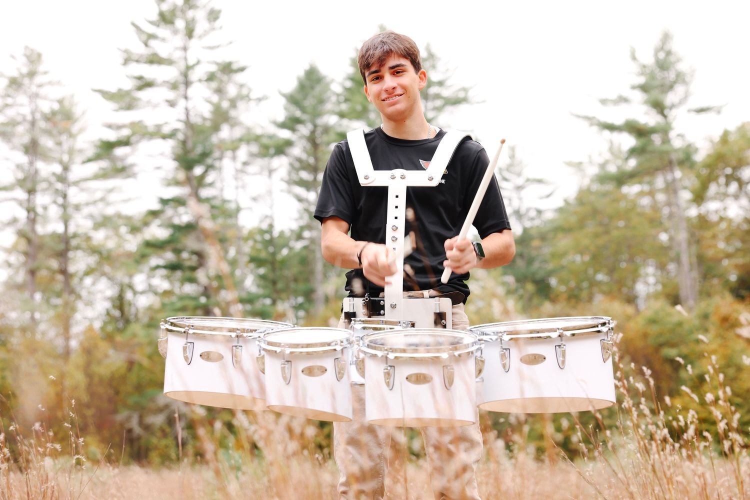 Young male drummer playing snare drums outdoors in a field surrounded by autumn trees.