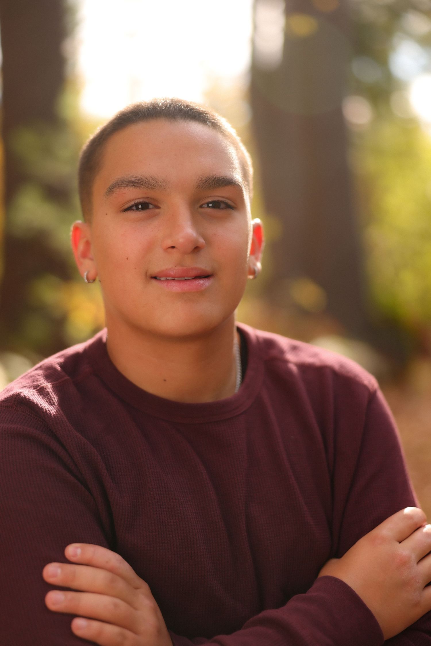 Smiling teenage boy with crossed arms posing outdoors in a maroon sweater in autumn light.