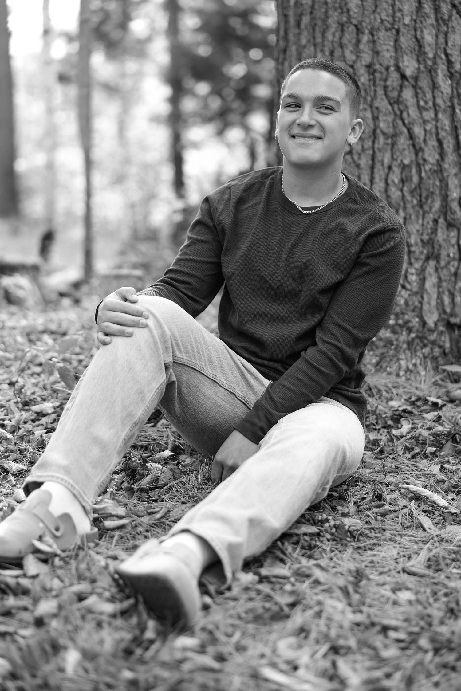 Black and white portrait of a young man sitting against a tree in a forest setting.