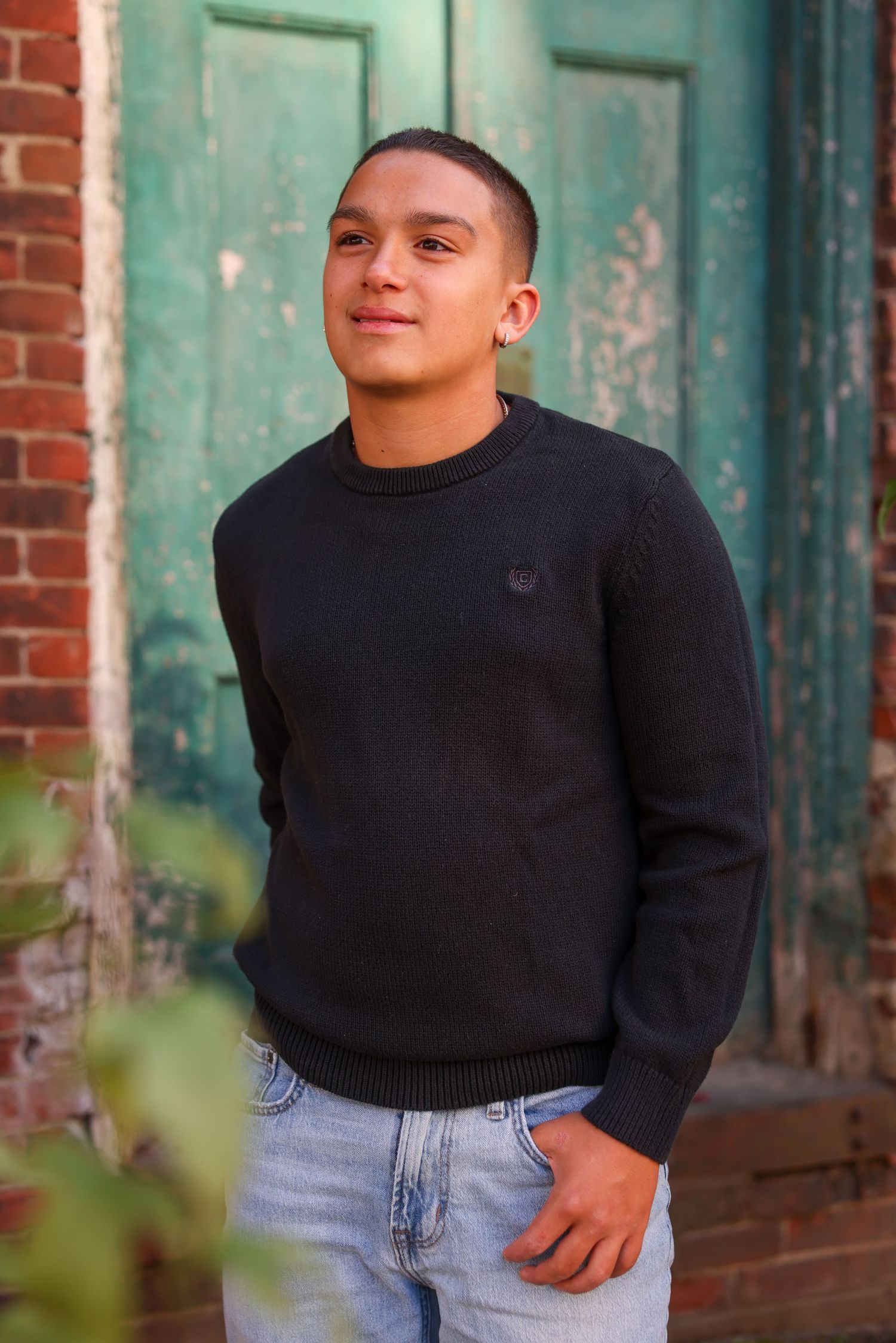 Young man in black sweater and jeans posing confidently by a rustic green door and brick wall.